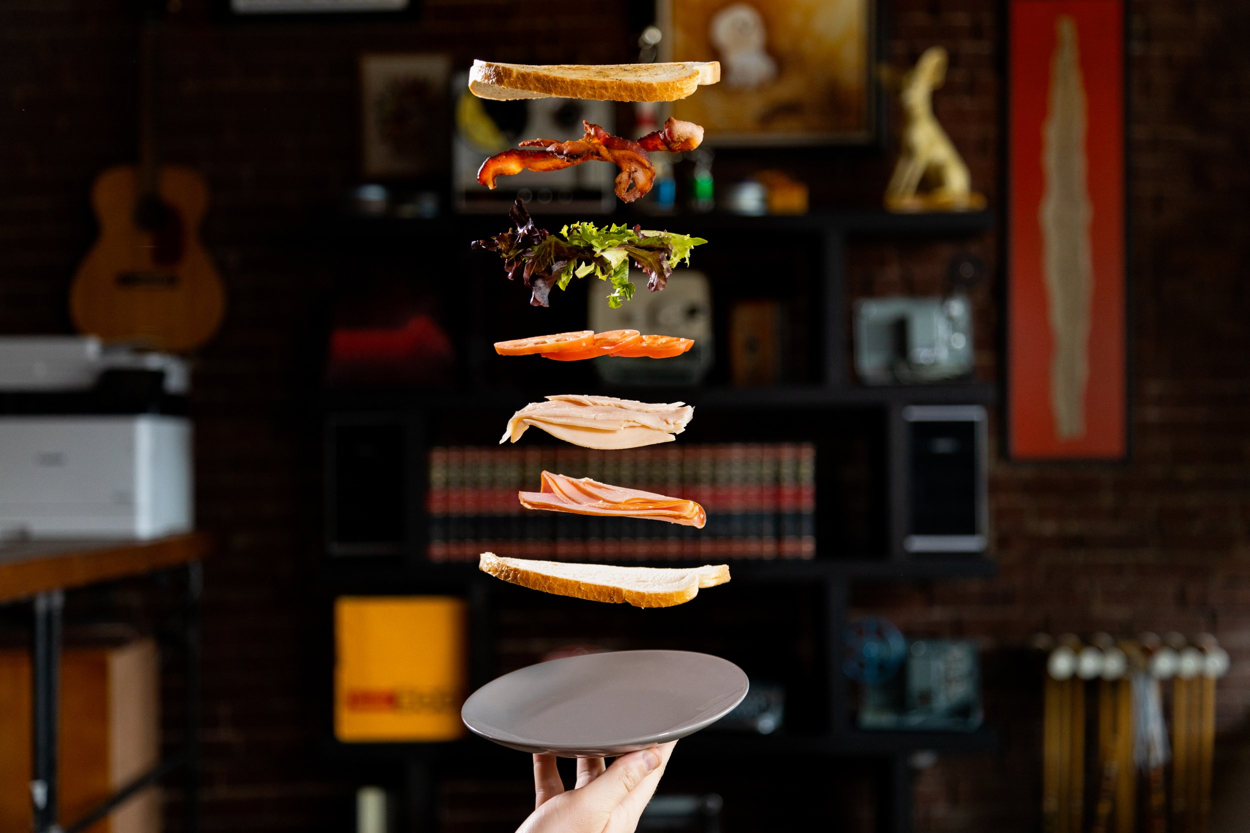 Floating sandwich ingredients including bread, bacon, lettuce, tomato, turkey, and ham over a plate in a kitchen setting.