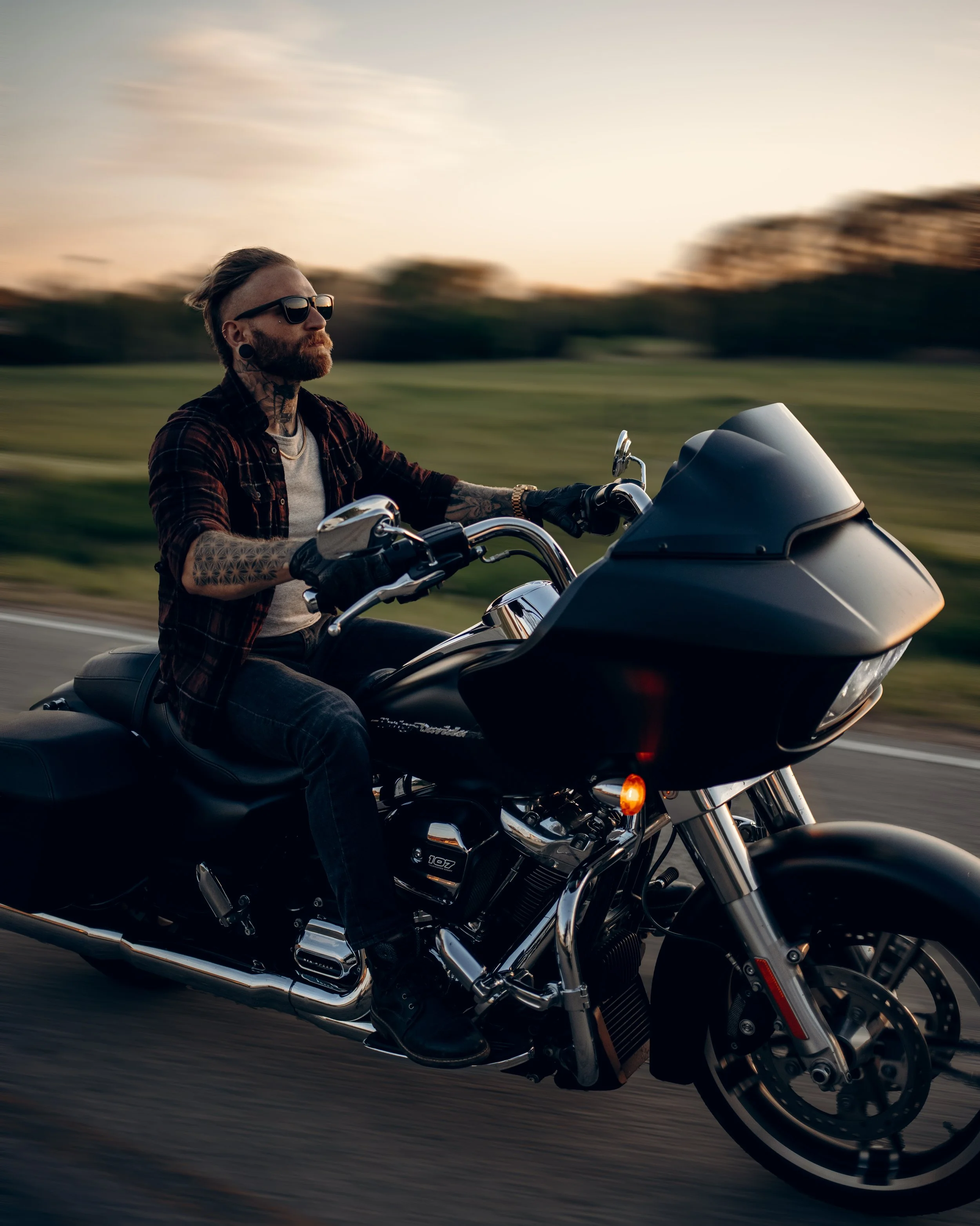 Man with tattoos riding a Harley-Davidson motorcycle on a road at sunset.
