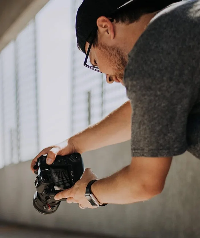 A man wearing glasses, a black cap, and a gray shirt is holding a camera, looking down at it. He is wearing a smartwatch and is in an indoor setting with a bright, grid-patterned window in the background.