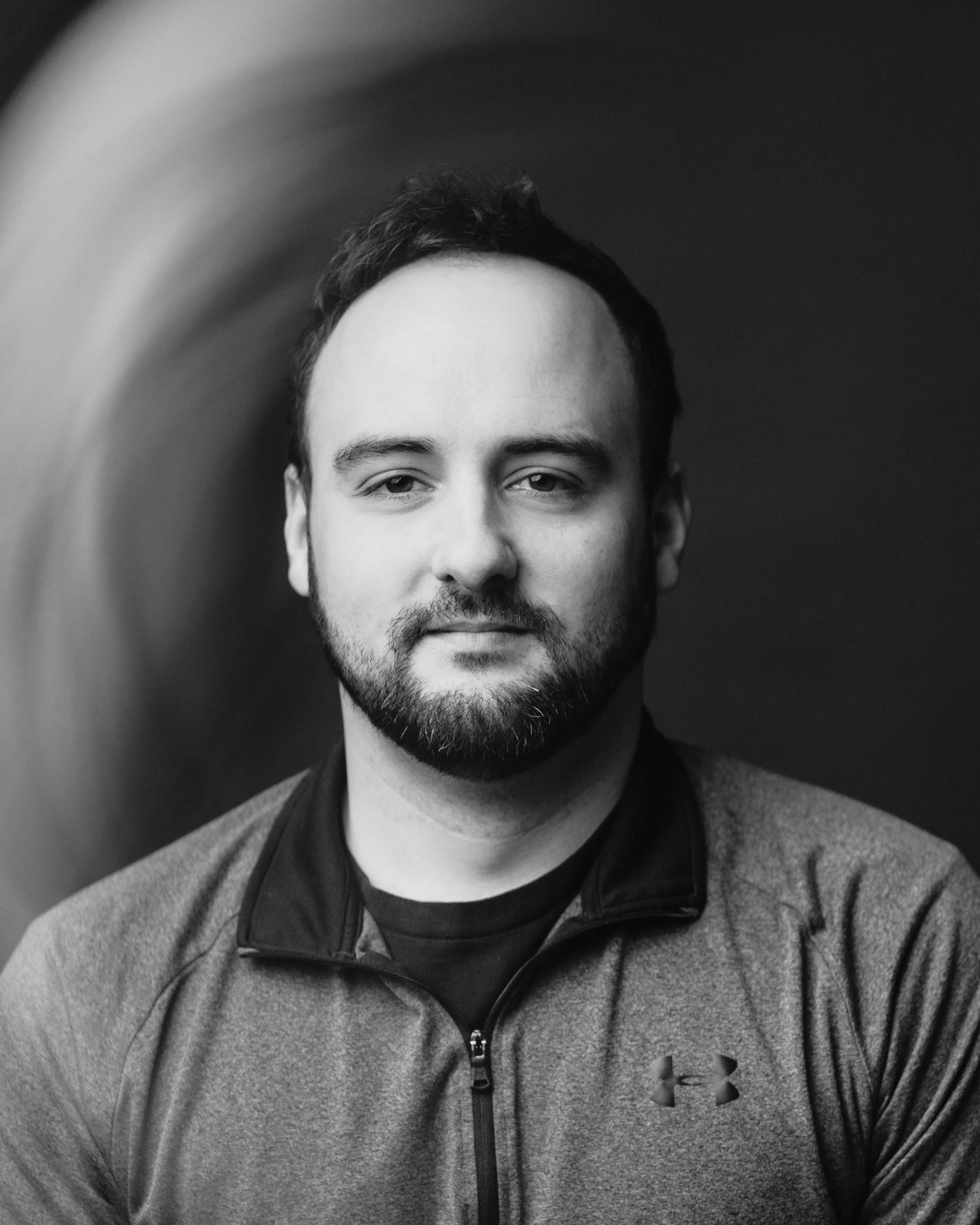 A black and white portrait of a young man with short, dark hair and a beard, wearing a zip-up hoodie with a small logo on the chest, looking directly at the camera.