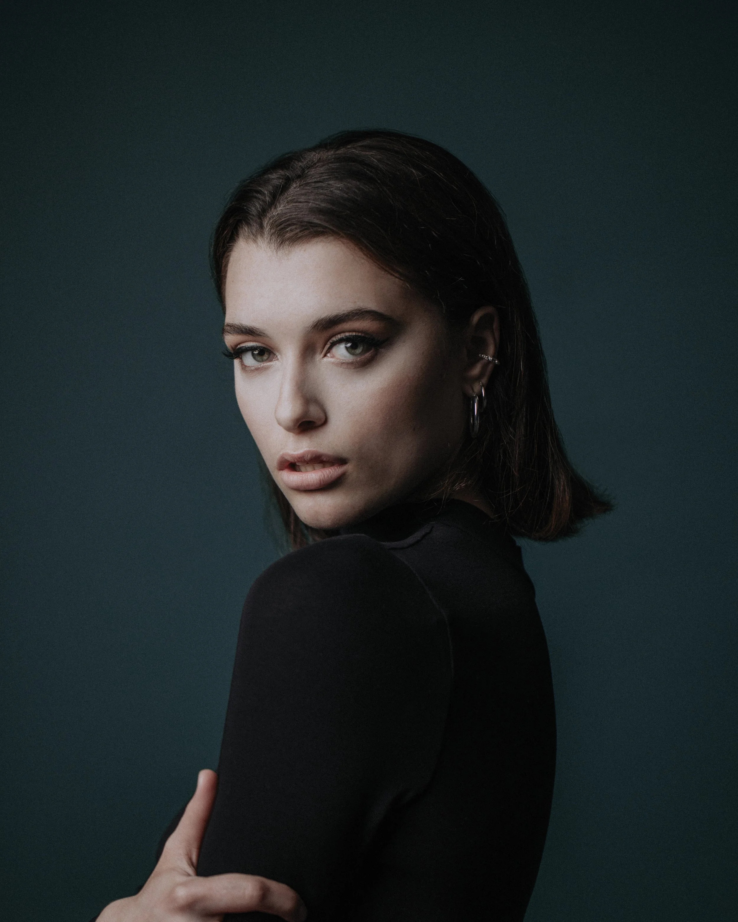 Woman in black top with short hair, looking over her shoulder, against a dark green background.