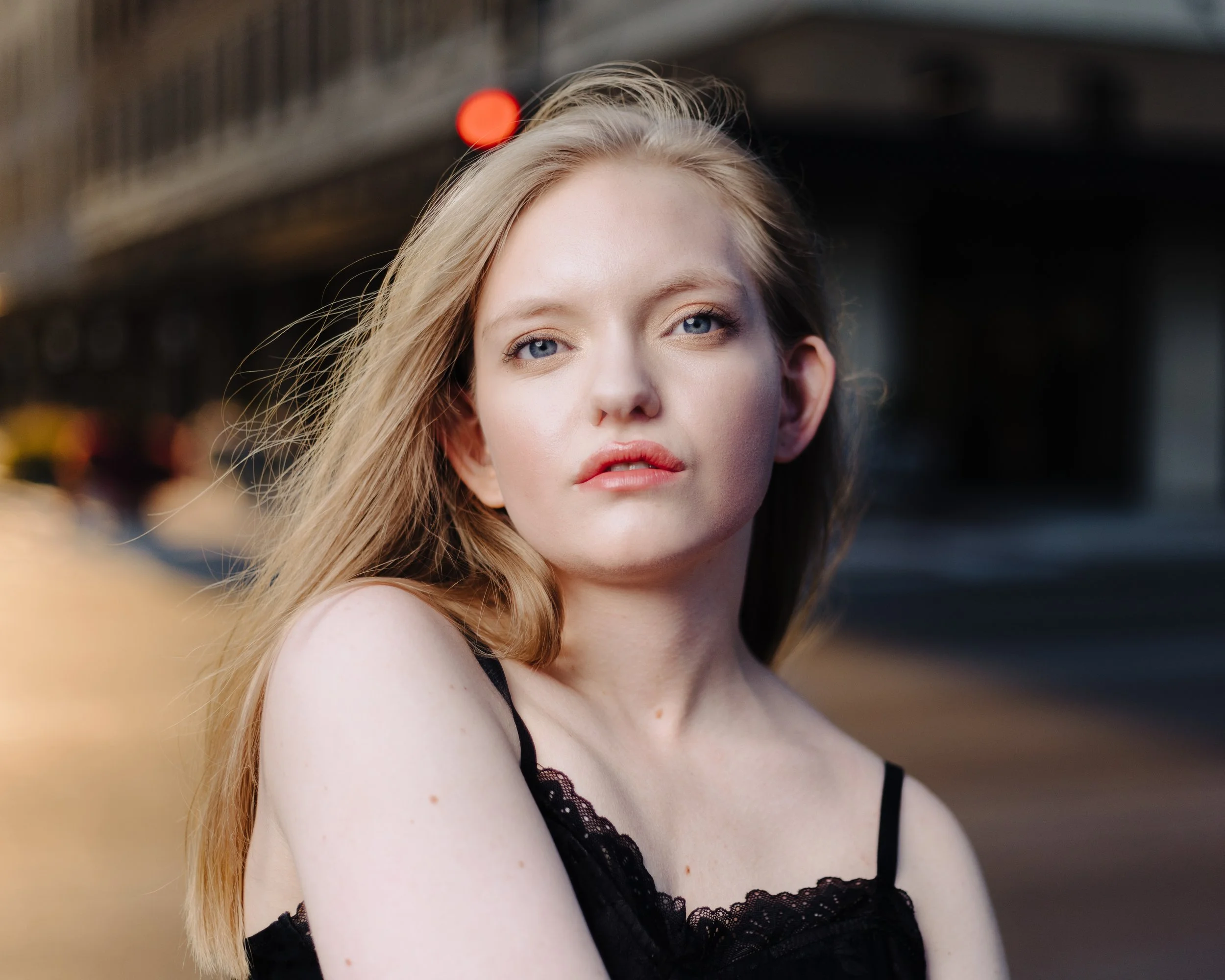Young woman with long blonde hair wearing a black lace top in an urban setting with blurred background and glowing streetlight.