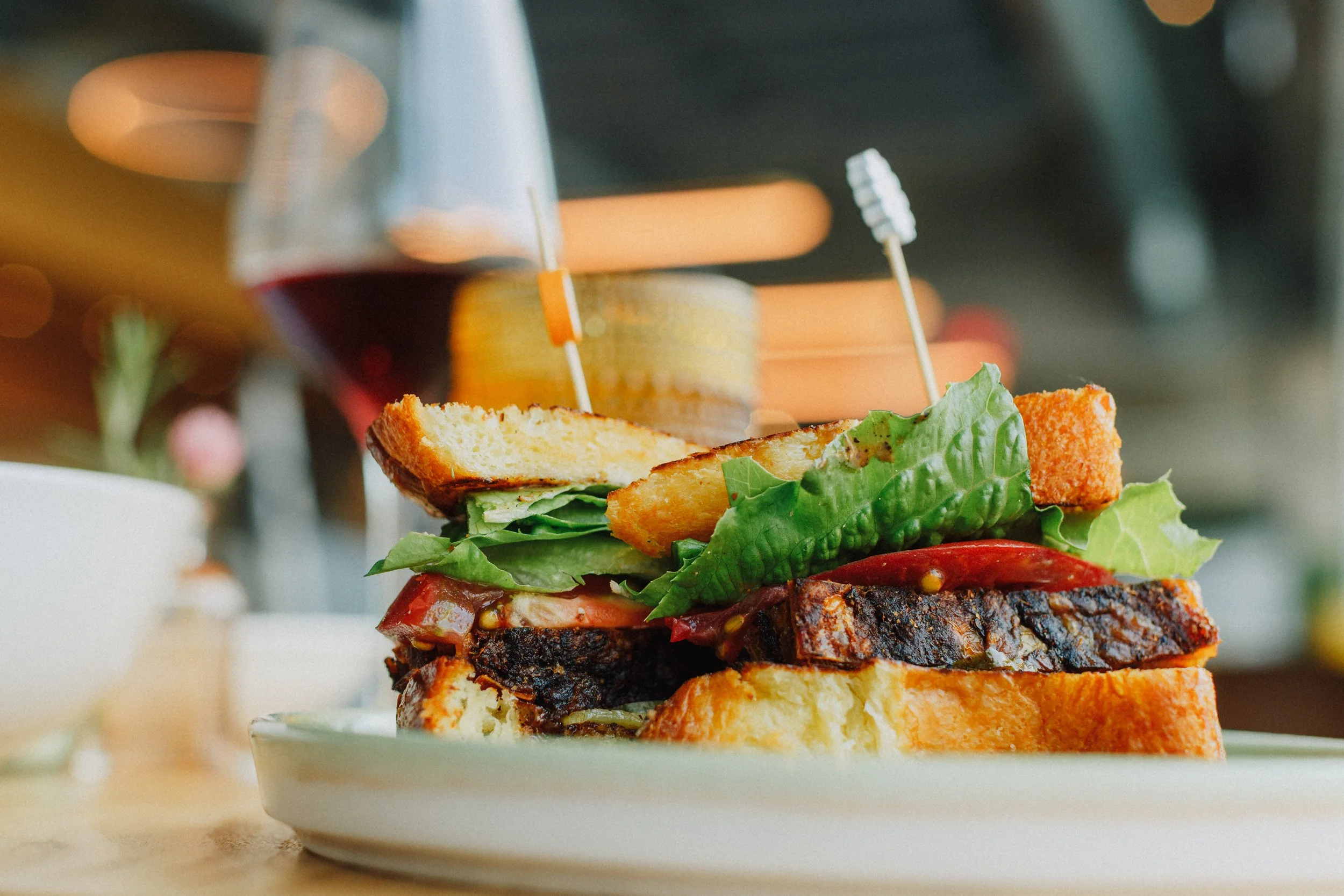 Close-up of a grilled sandwich with lettuce, tomato, and fried chicken, served on a white plate, with a glass of red wine in the background.