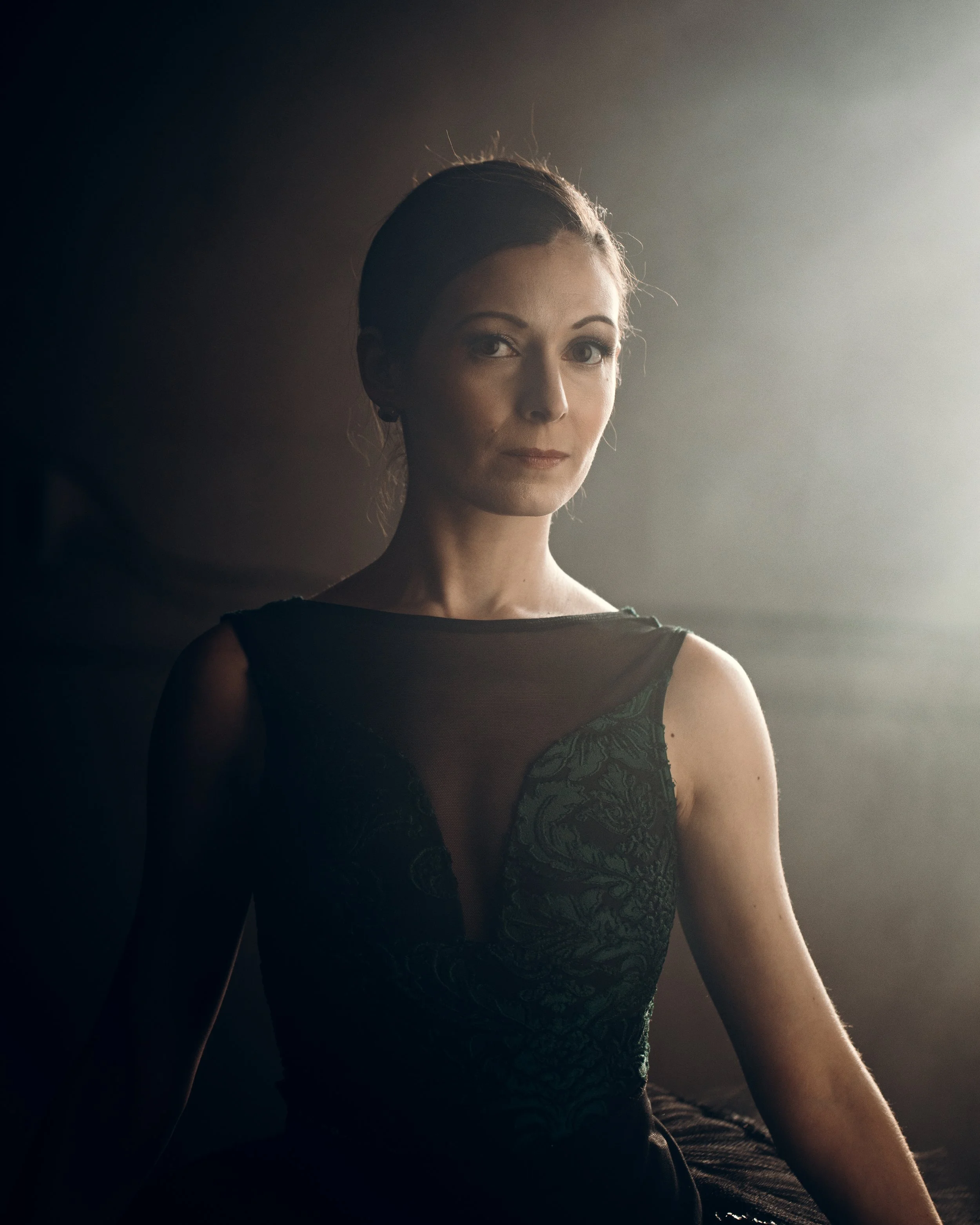 Portrait of a woman in an elegant dress, sitting, with a neutral expression, softly lit with a dark background.