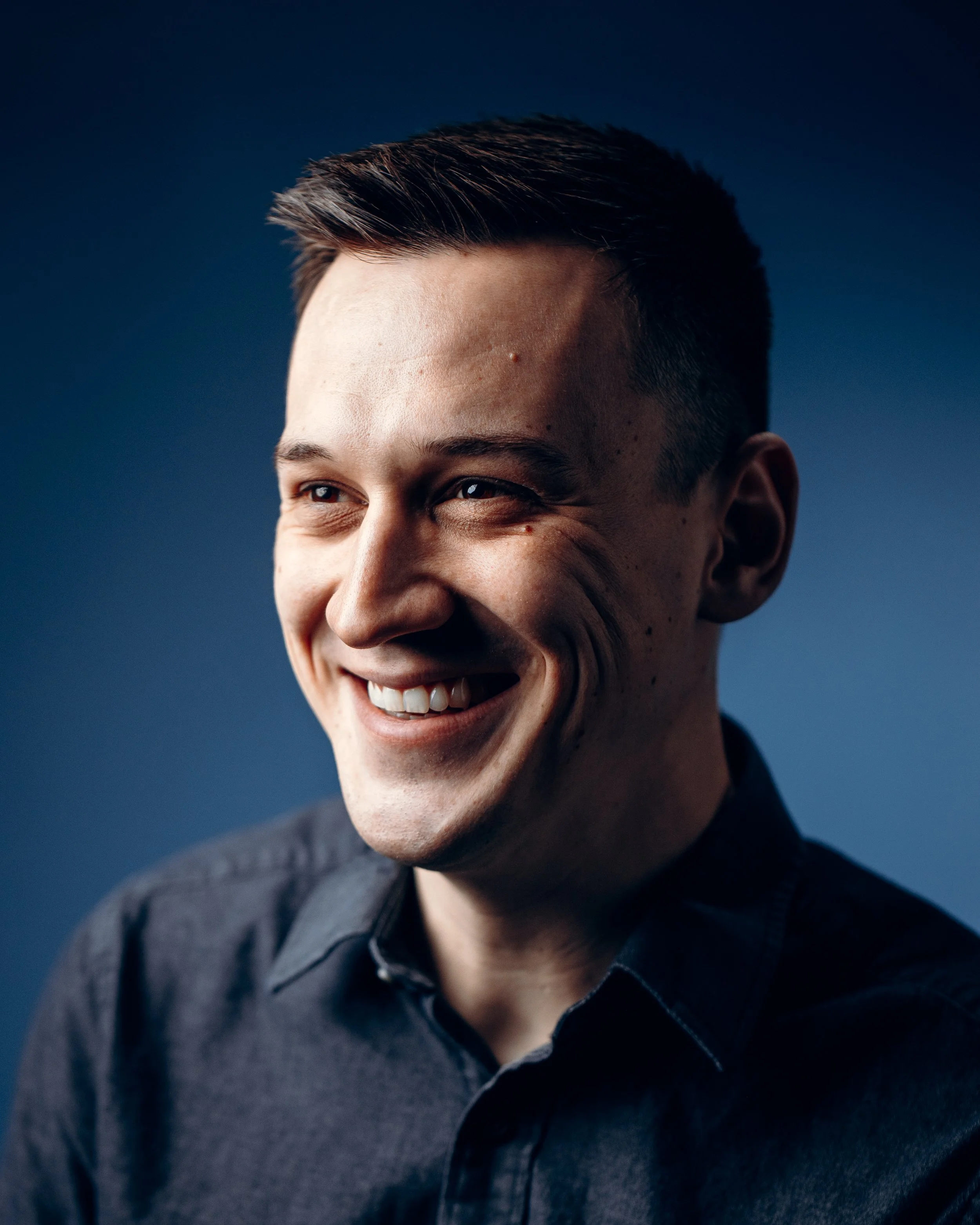 Smiling man in a dark shirt, looking to the side, with a dark blue background.
