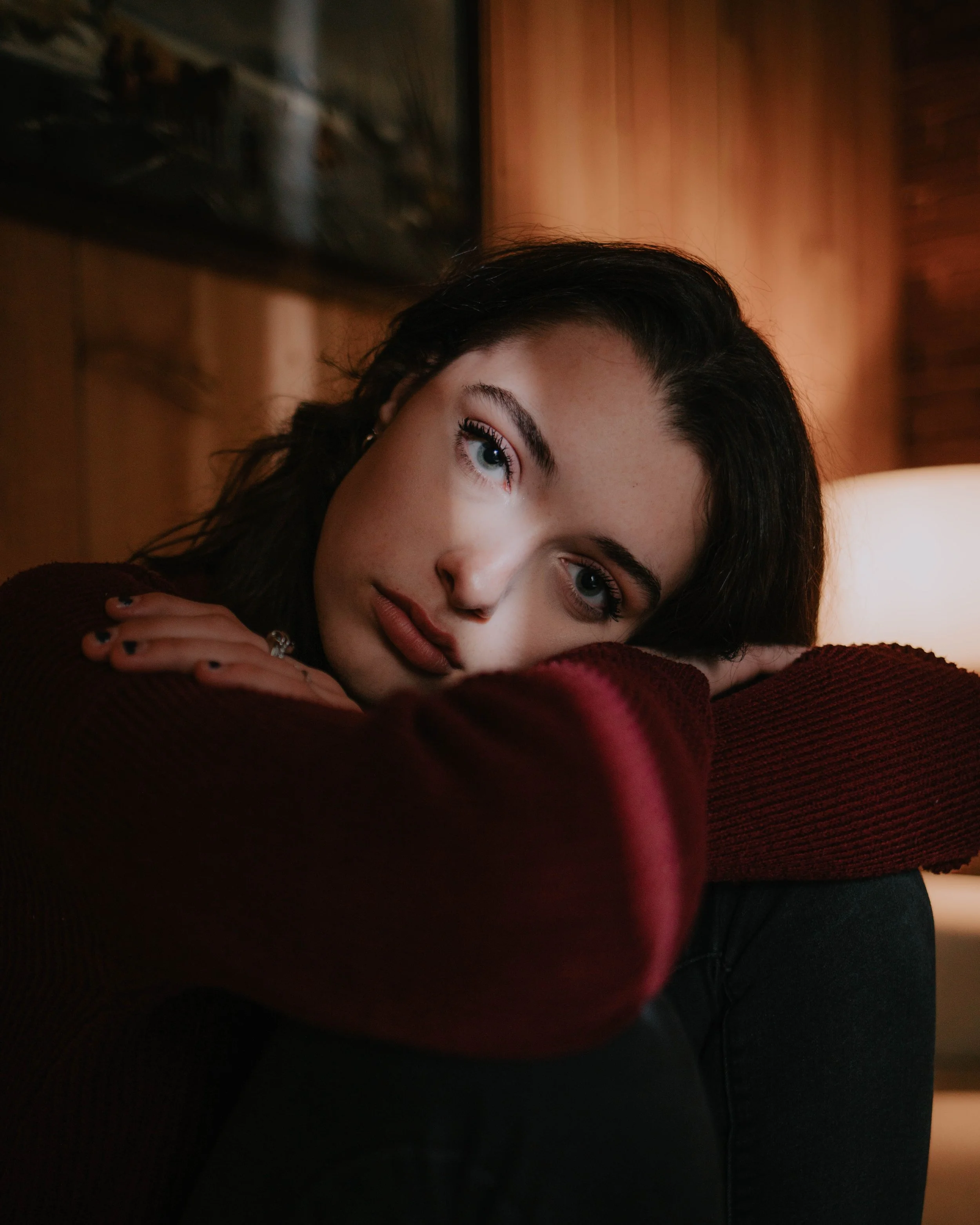 Woman in a dimly lit room gazing contemplatively, resting her head on folded arms, wearing a dark maroon sweater.