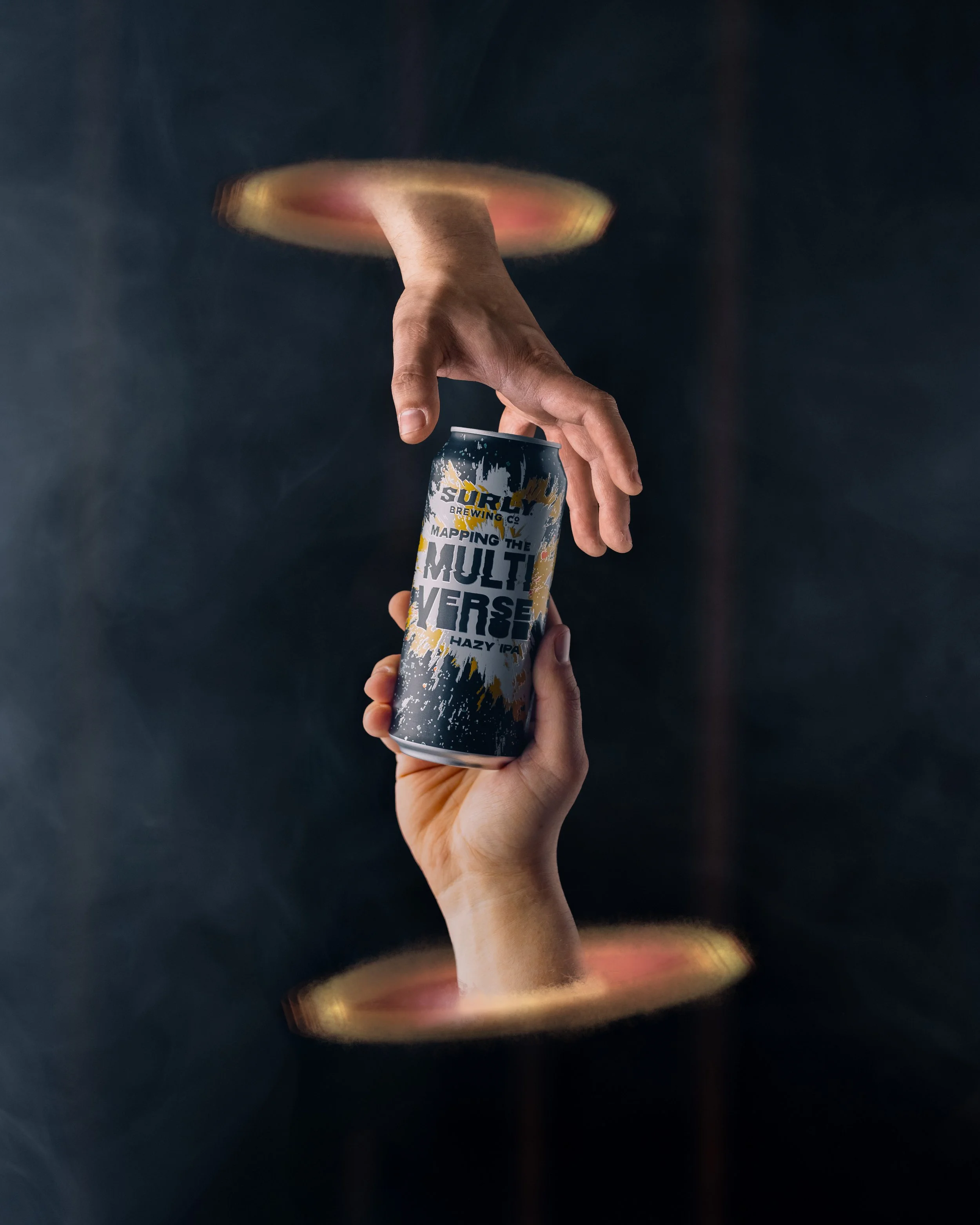 Two hands, one receiving a beer can labeled 'Mapping the Multiverse Hazy IPA,' emerging from portal-like rings against a dark background.