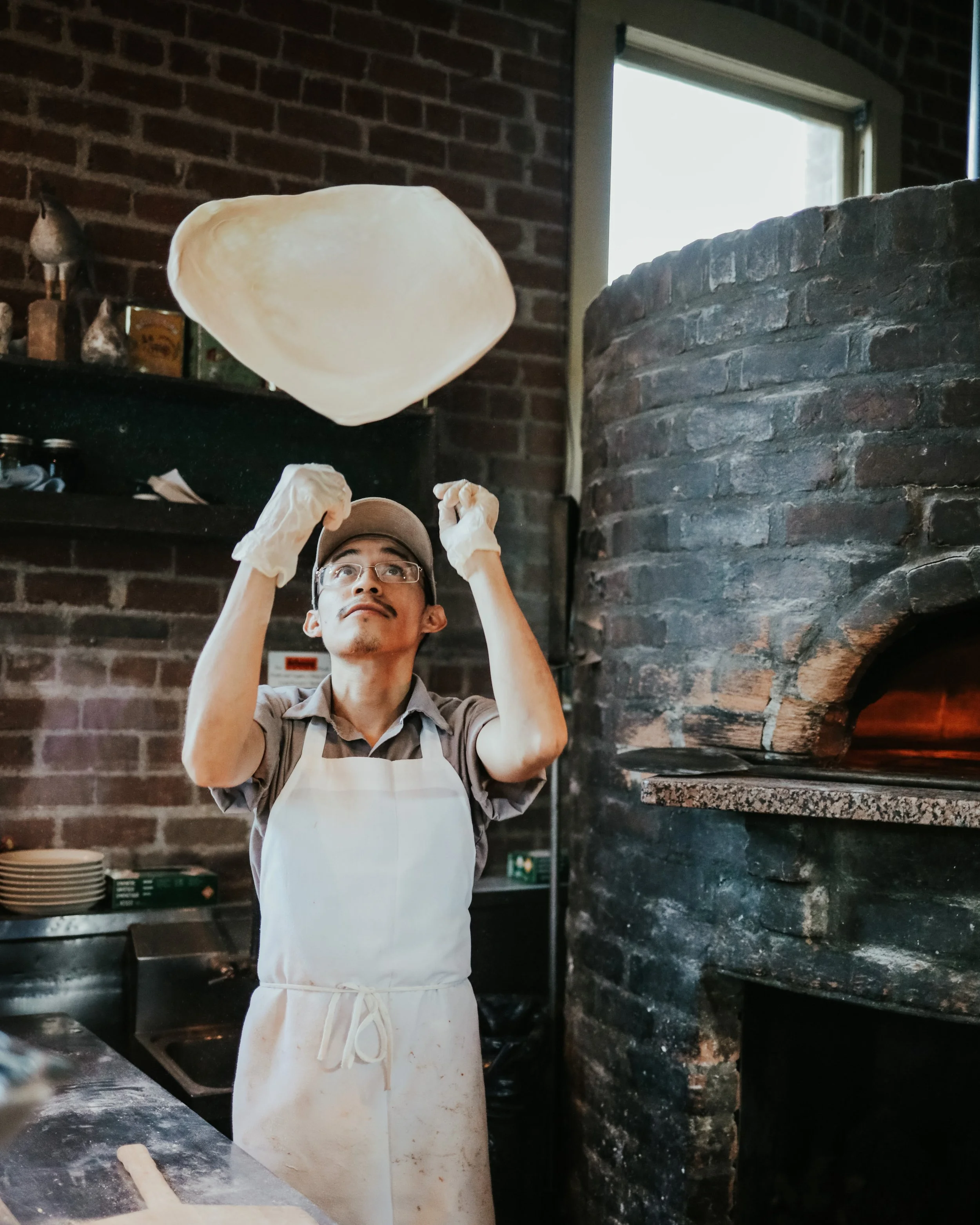 Pizza chef tossing dough near a brick oven in a rustic kitchen.