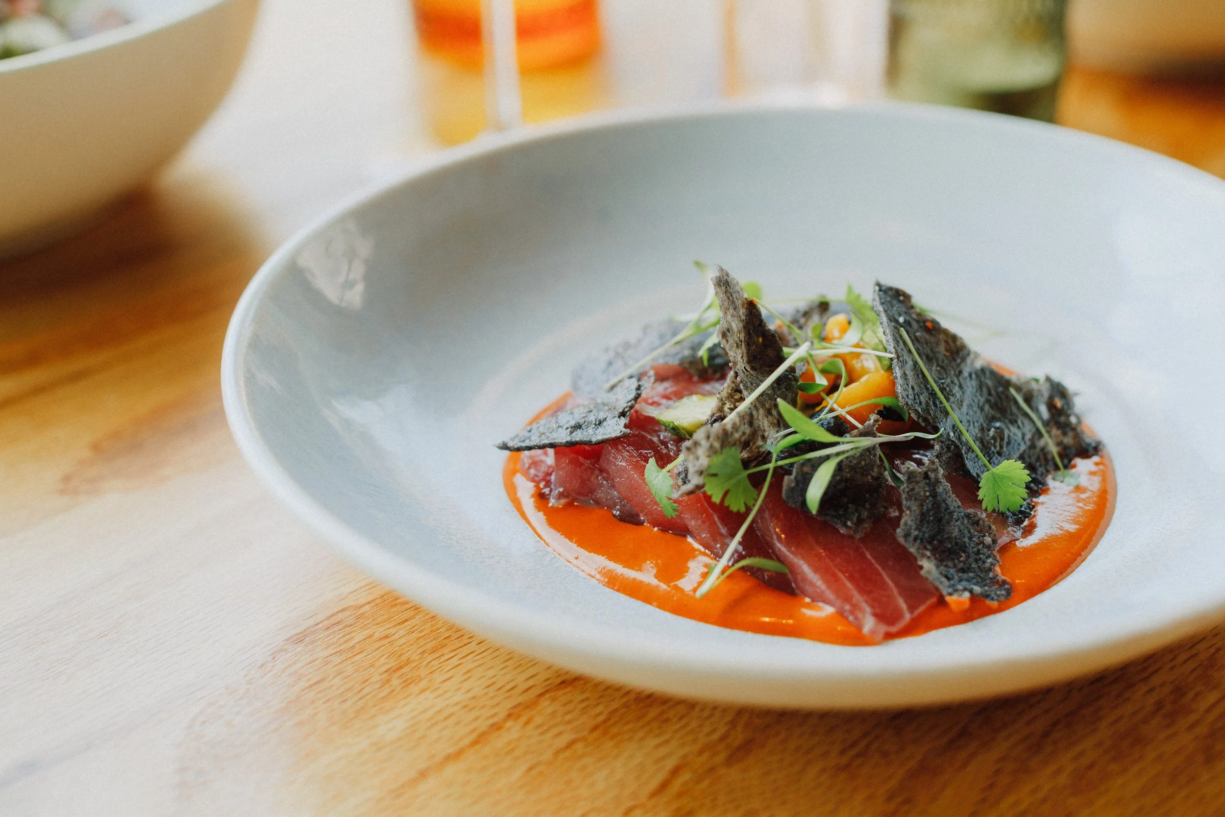 A plate of seared tuna with black crisps, microgreens, and a red sauce on a wooden table.