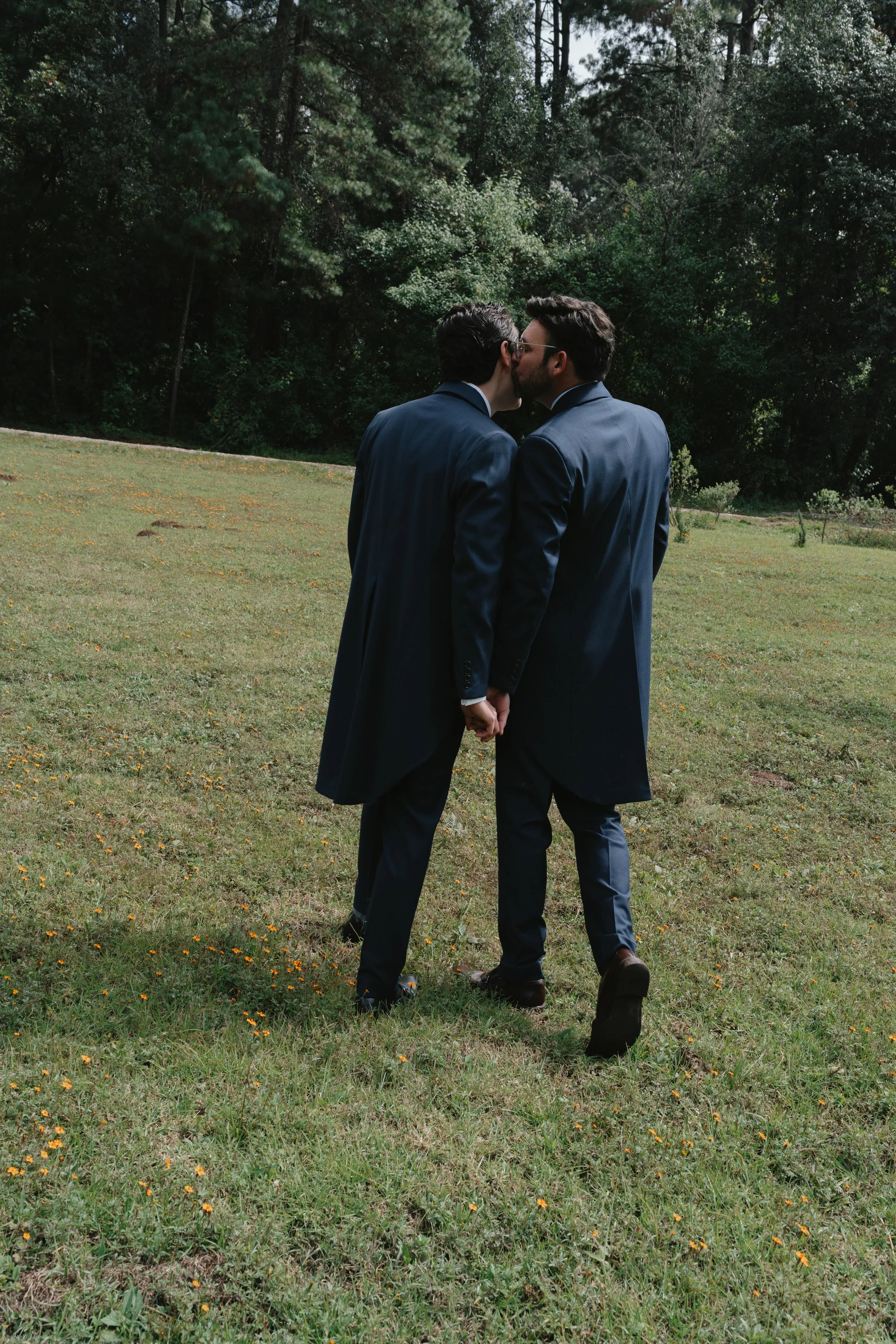 Dos hombres vestidos con trajes oscuros y abrigos se besan en un campo rodeado de árboles.