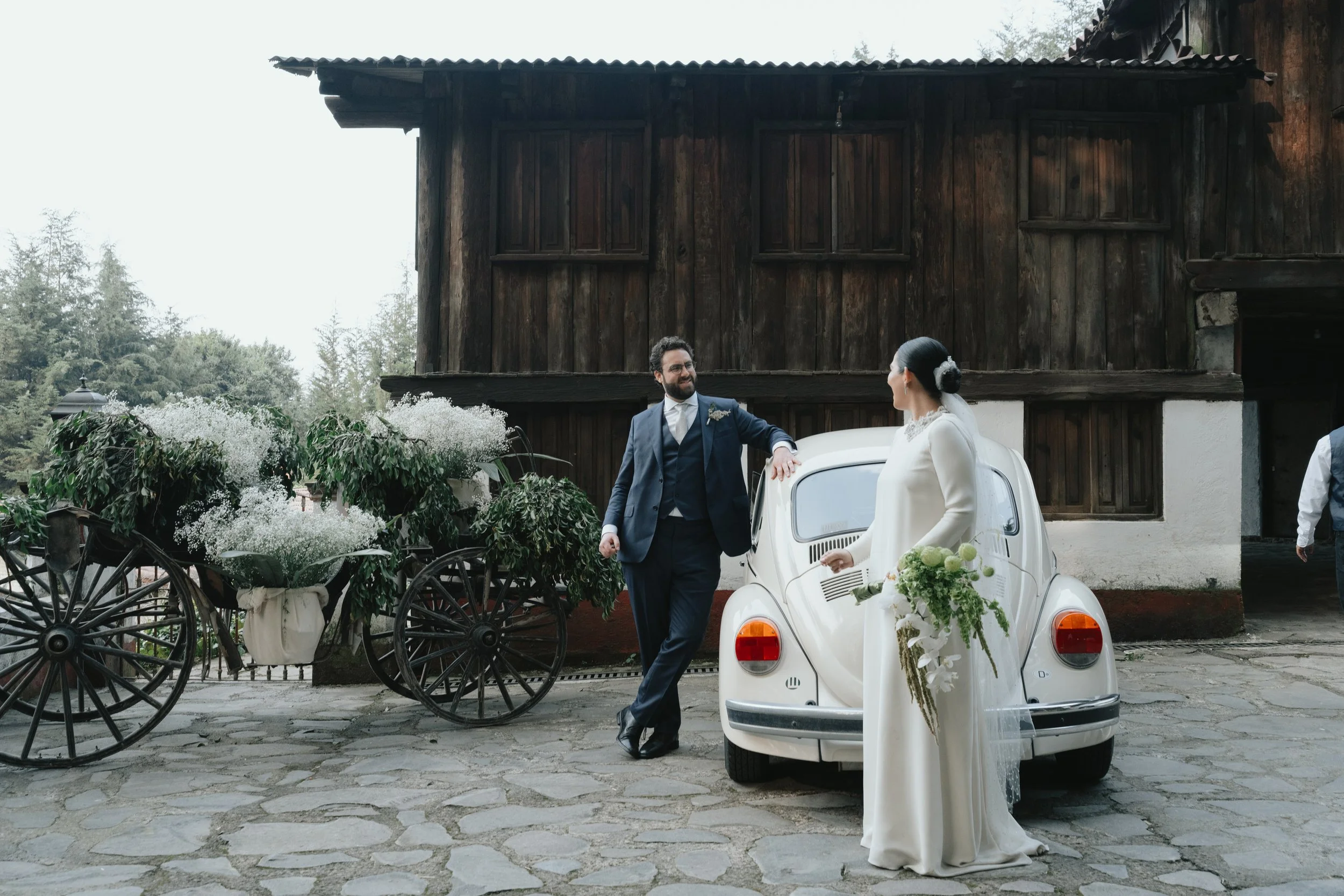 Pareja de novios en boda, el novio de traje azul y la novia de vestido blanco, conversando junto a un coche clásico blanco y una carreta decorada con flores blancas, en un entorno rural con casa de madera detrás.