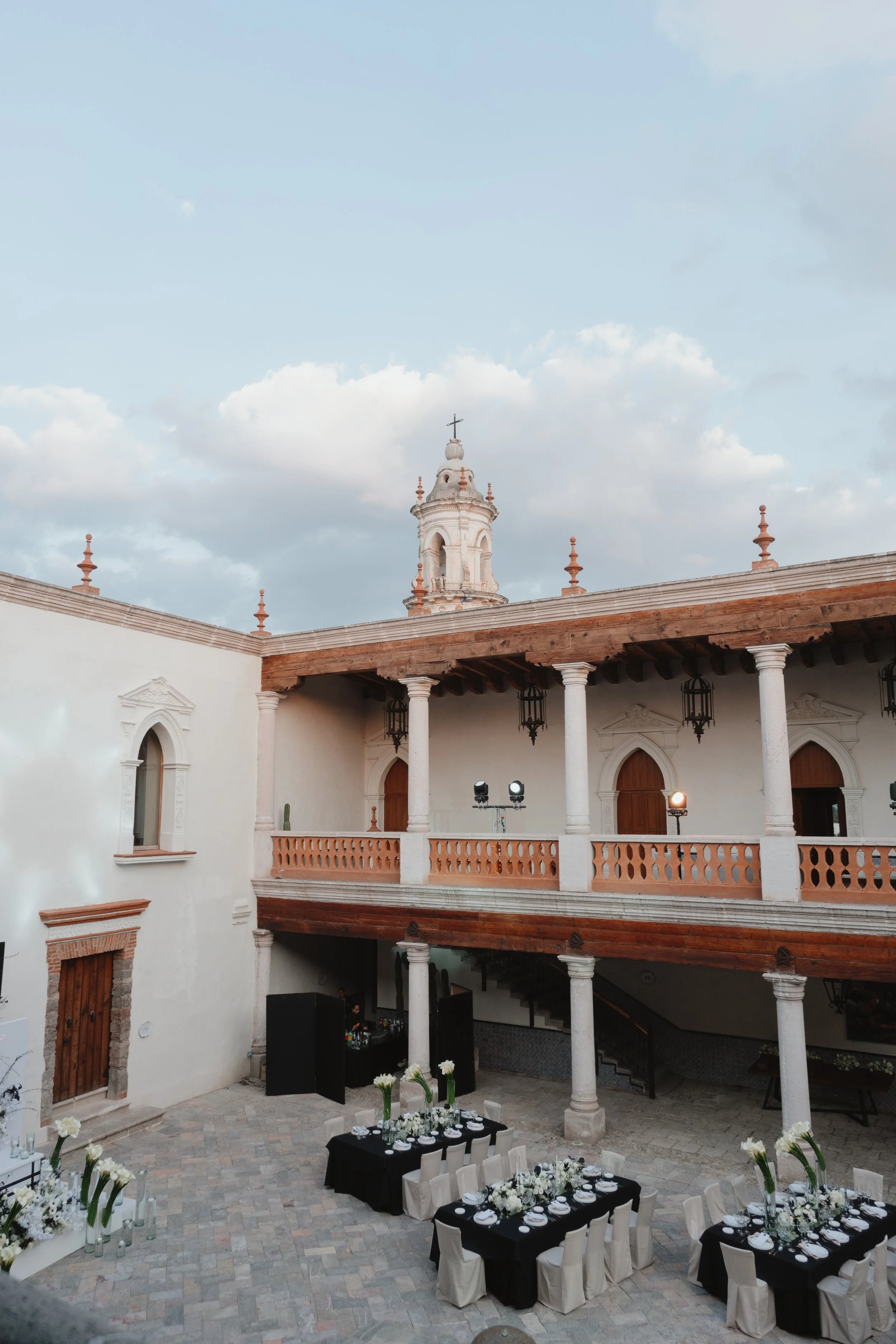 Escenario para una celebración en un patio interior con mesas decoradas con flores blancas y un fondo de arquitectura colonial con columnas y un campanario.