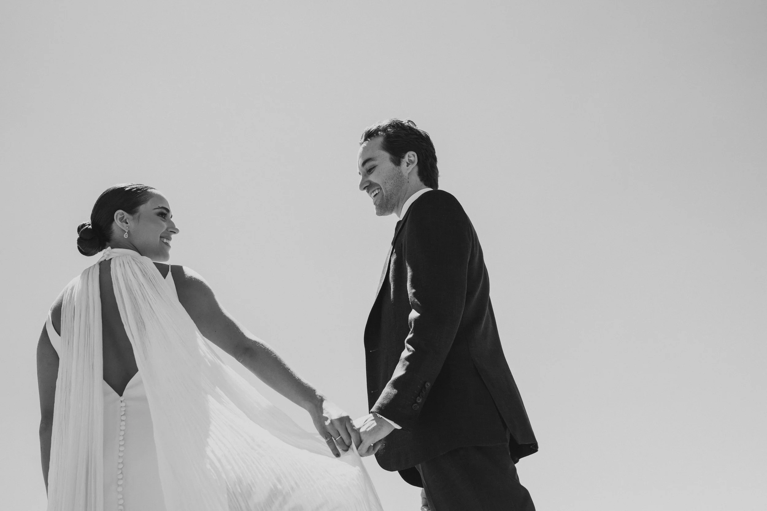 Pareja de recién casados en su boda, tomados de las manos y sonriendo en un fondo de cielo despejado.