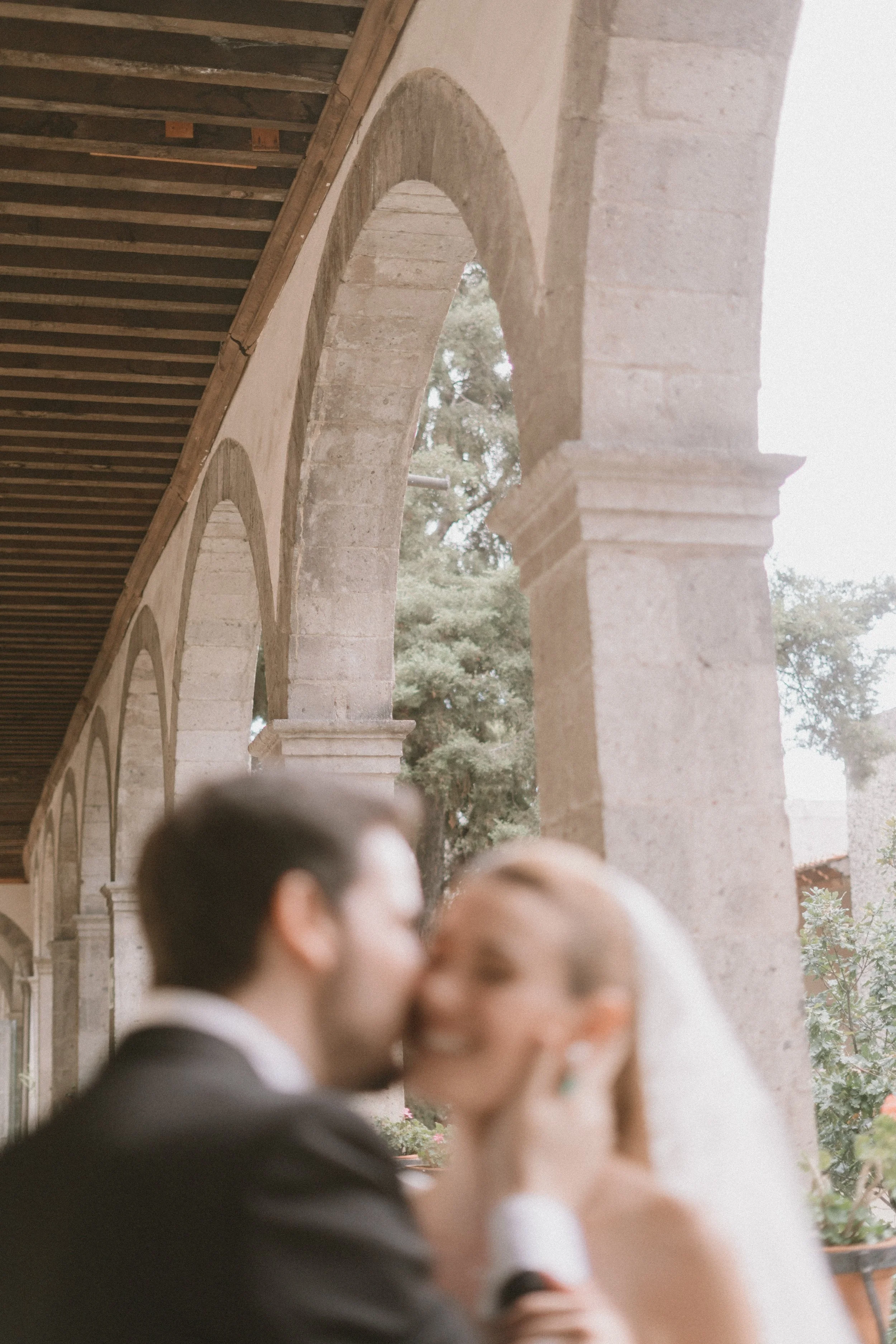 Boda en un lugar colonial con arcos de piedra y vegetación en el fondo, pareja de novios en primer plano, la imagen está desenfocada.