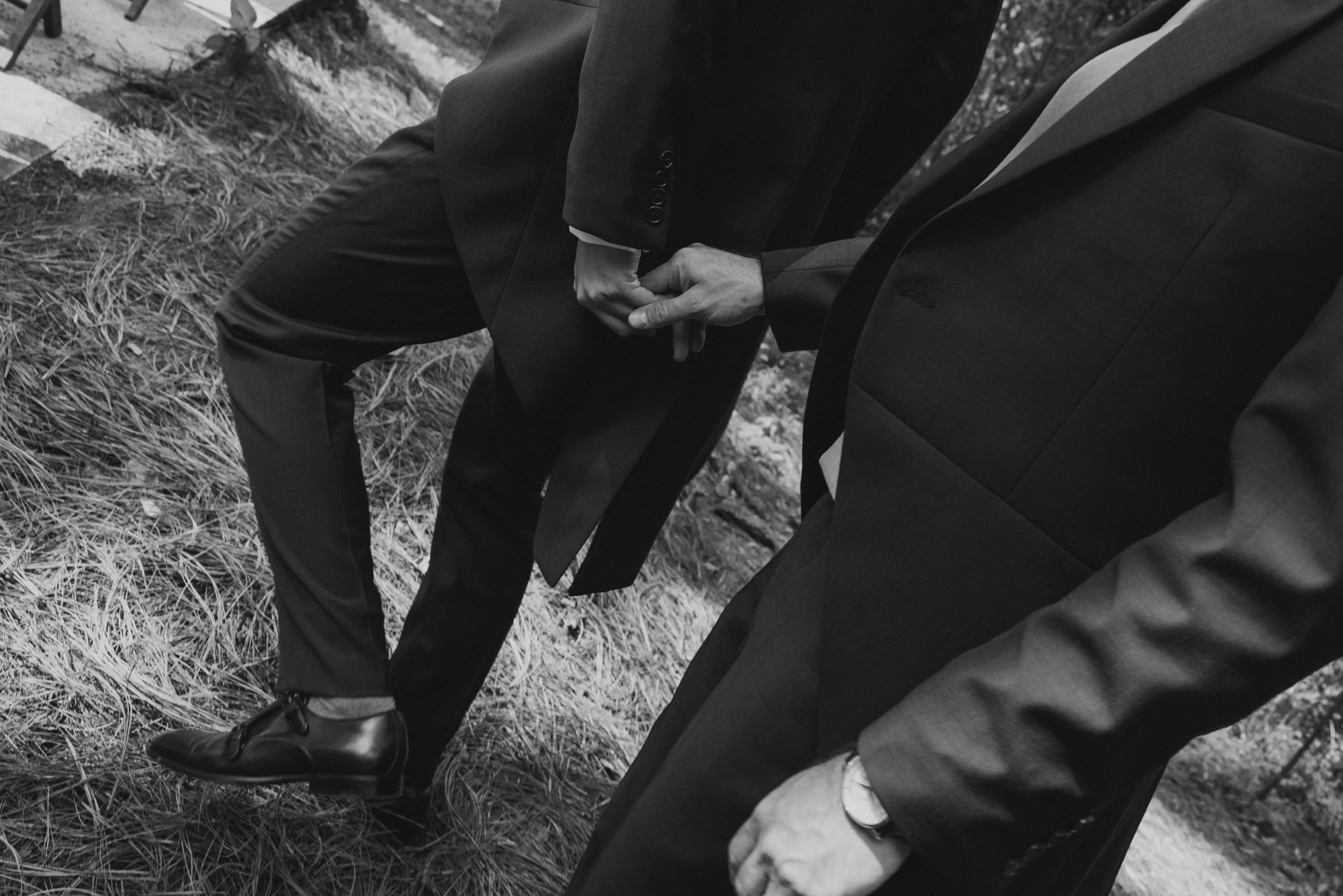 Dos hombres en trajes formales tomados de la mano en un entorno natural.
