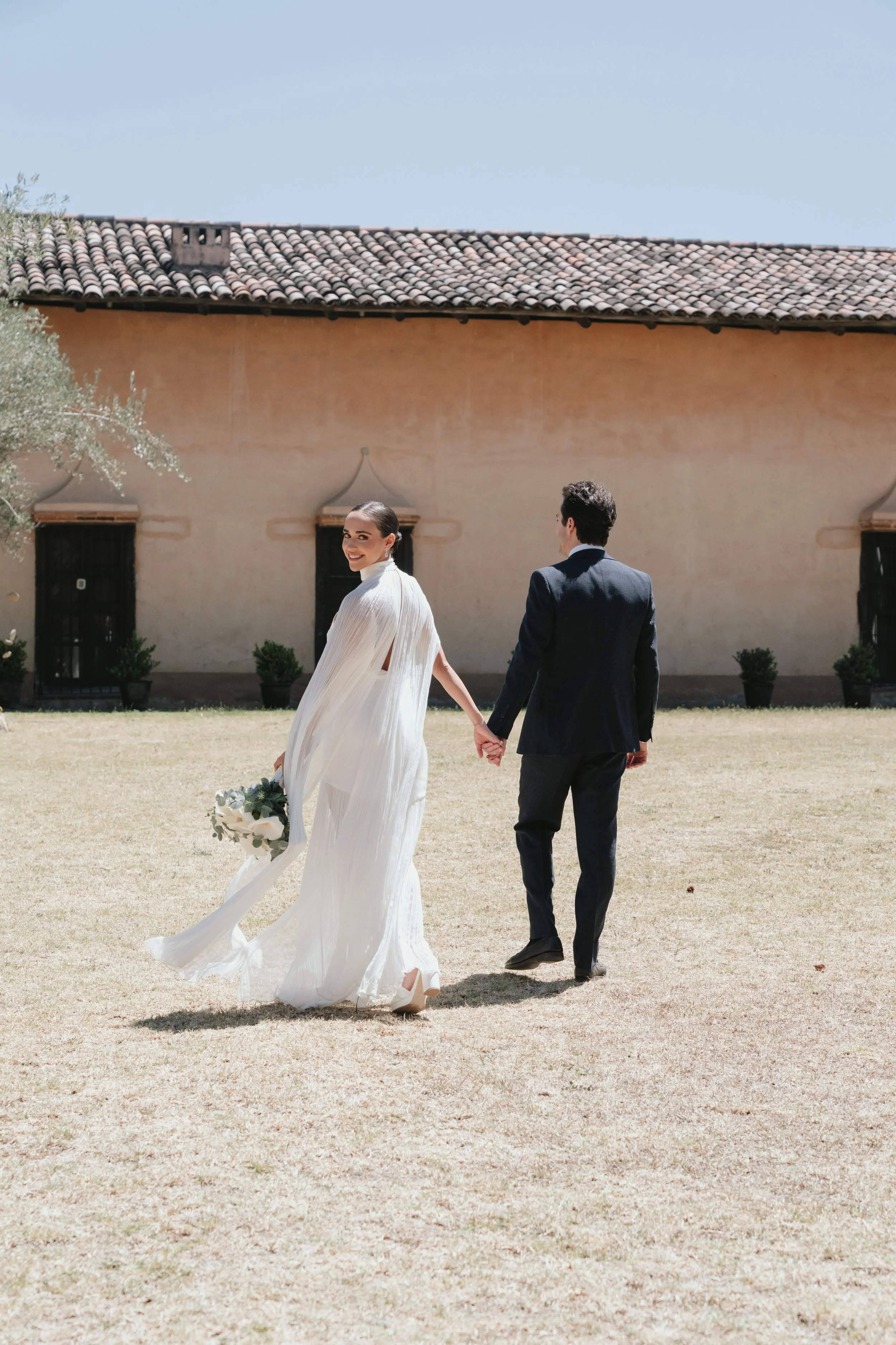 Pareja de novios caminando en un jardín, la novia sonriente con vestido blanco y ramo, el novio con traje oscuro, fondo de casa con tejas y pared de color beige.