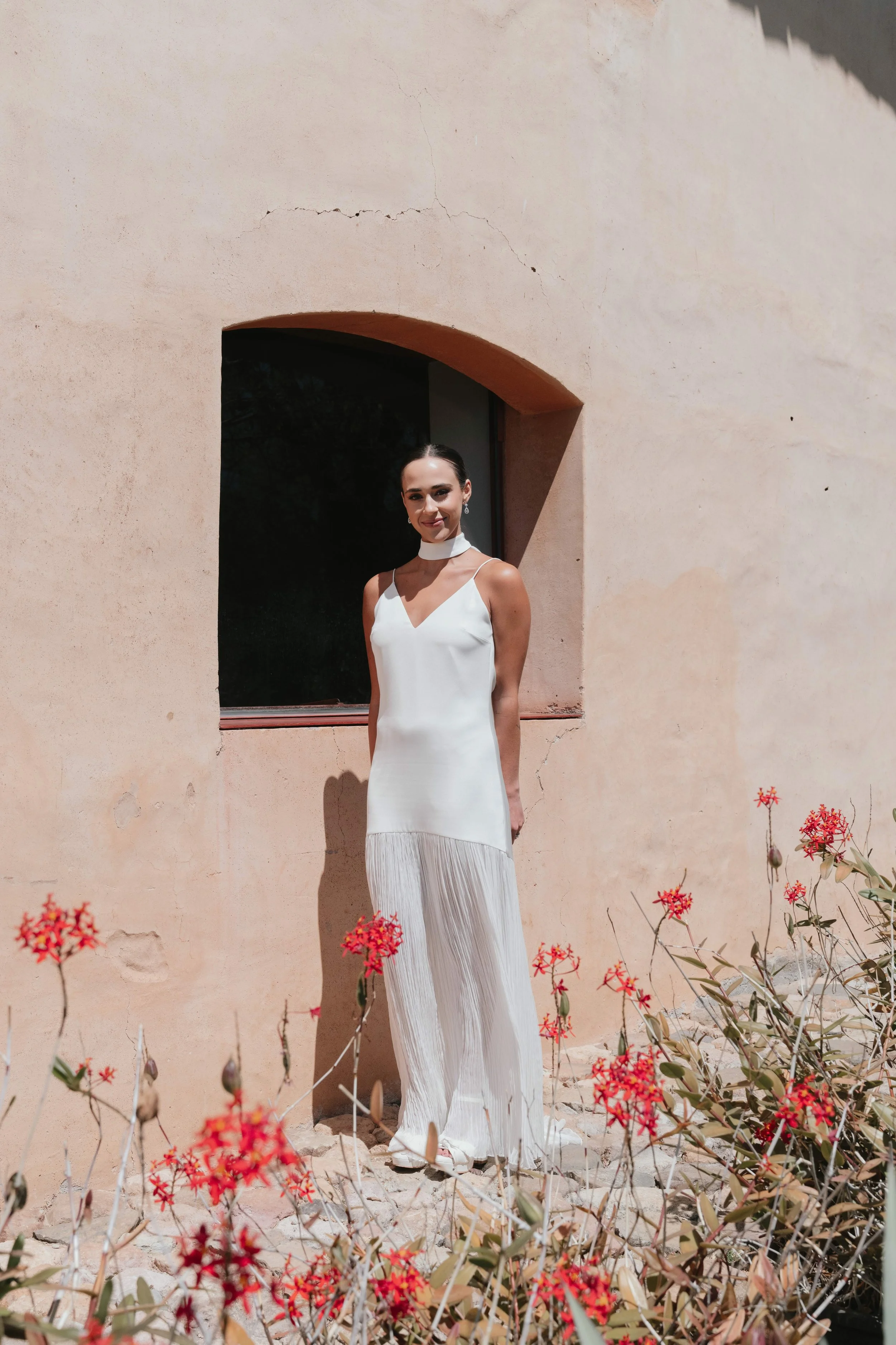 Mujer de pie junto a pared de color beige con flores rojas y verdes en primer plano, lleva vestido blanco largo y cuello alto, sonriente, en un entorno exterior soleado.