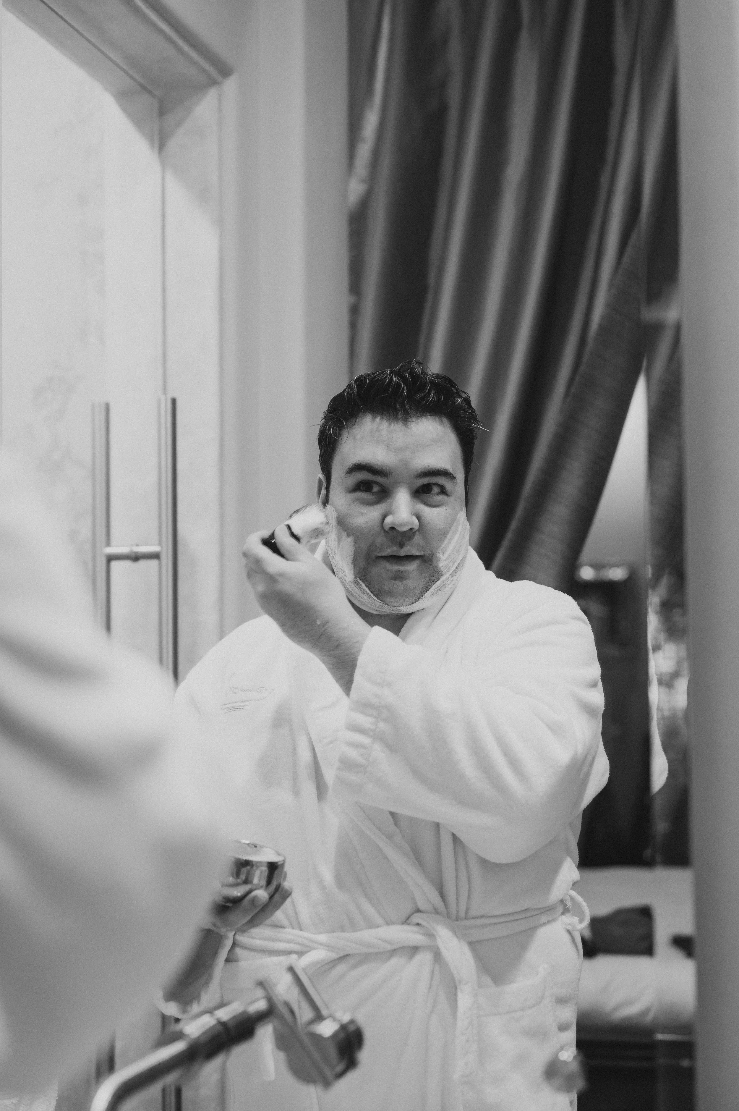 Hombre en bata de baño se afeita frente a un espejo en un baño decorado con cortinas elegantes.