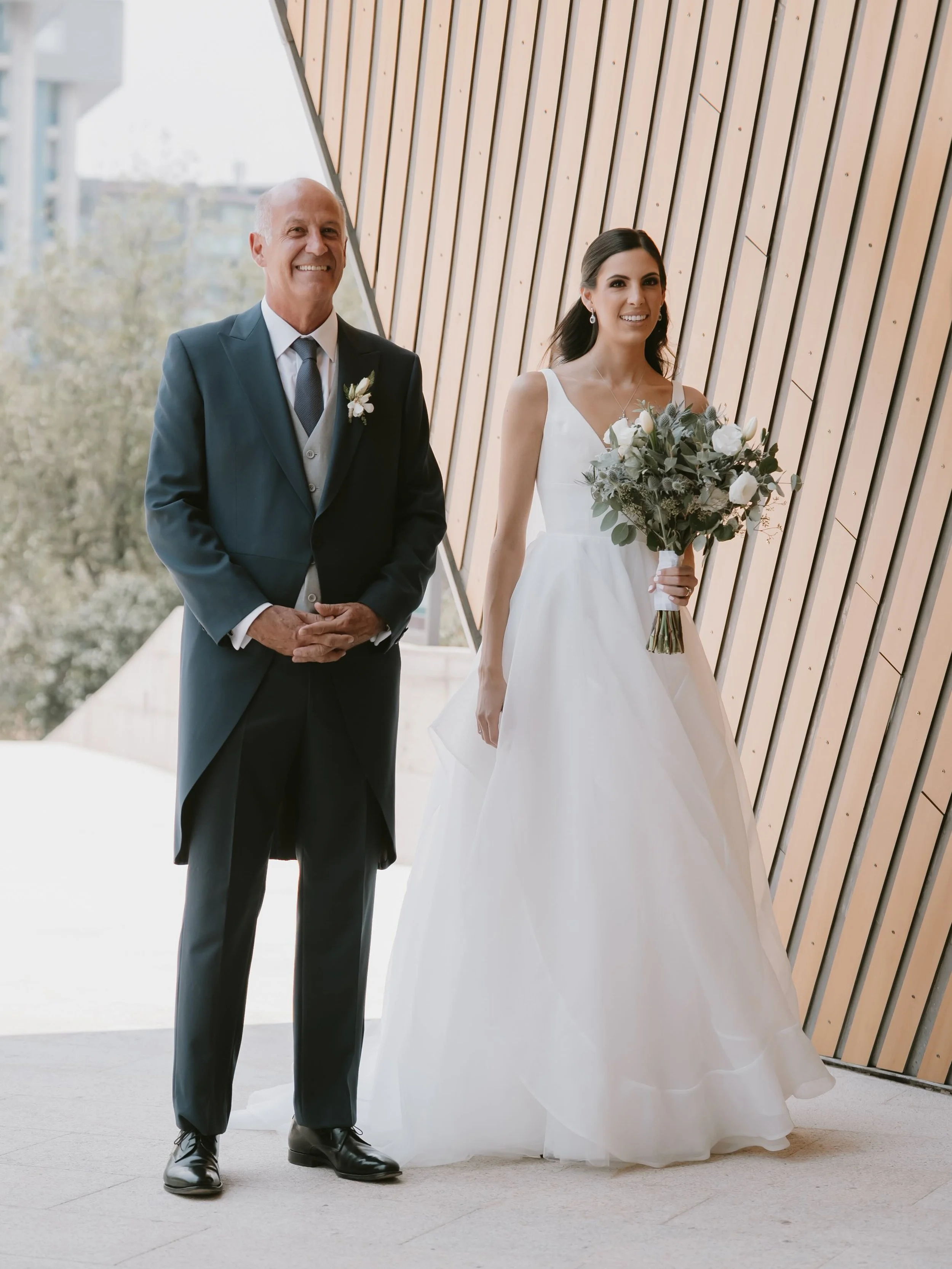 Mujer en vestido de novia con un ramo de flores, acompañada por un hombre en traje formal, en un lugar con pared de madera
