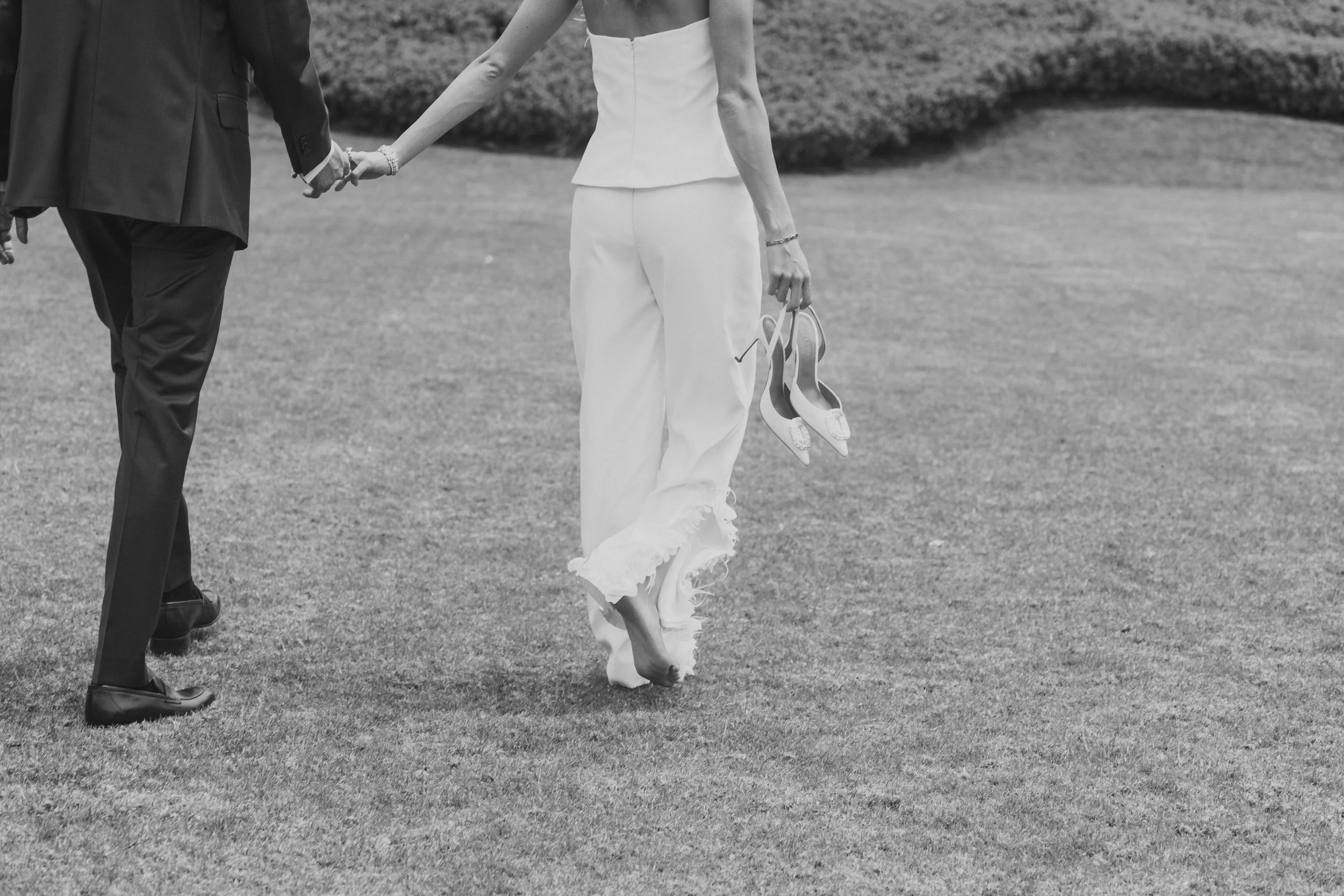 Pareja caminando en un campo, ella lleva zapatos y pantalones blancos, él lleva traje oscuro, en blanco y negro.
