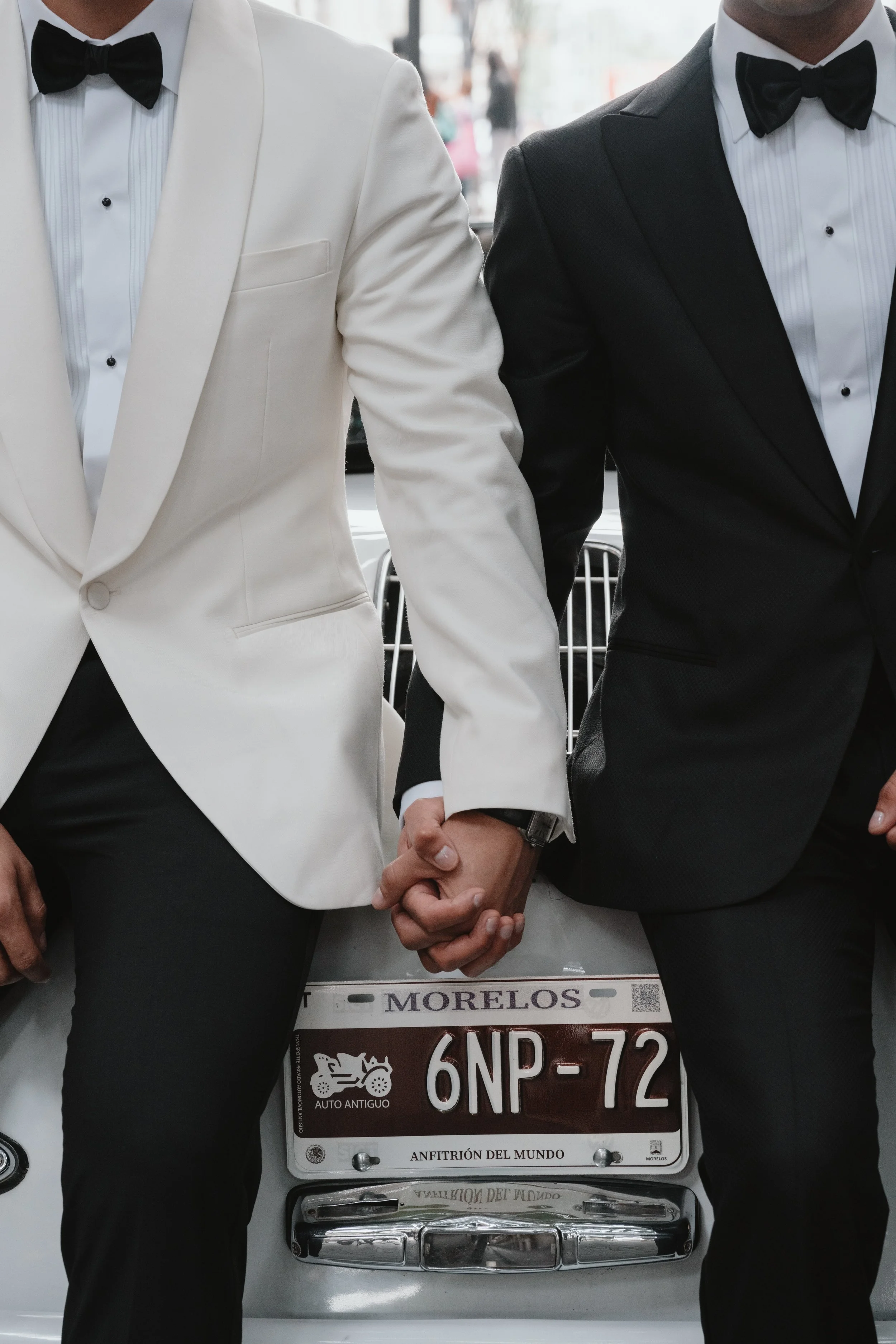 Dos hombres con trajes formales de esmoquin, uno blanco y otro negro, sosteniéndose de las manos y de fondo un auto con placa de Morelos, México.