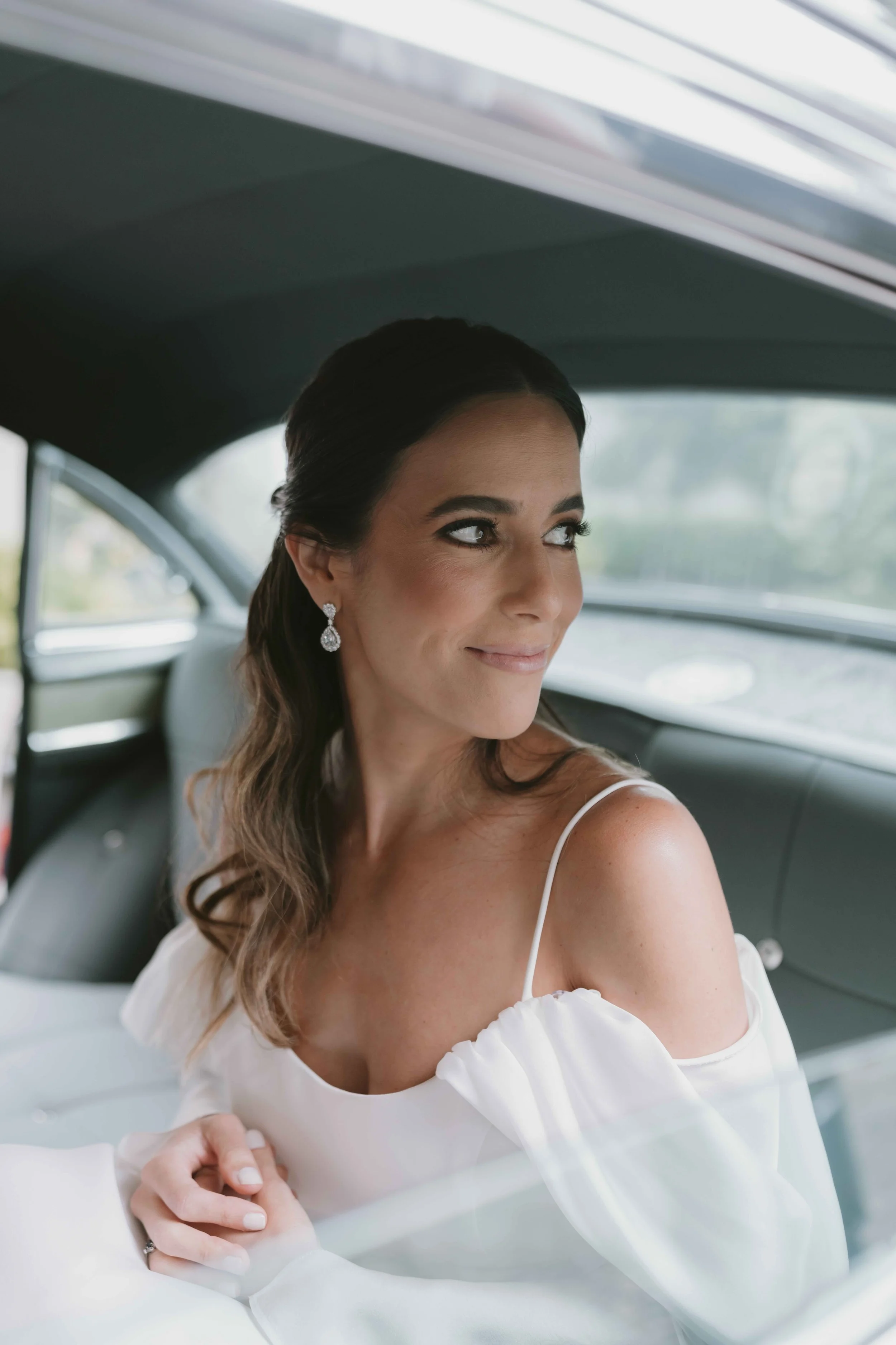 Mujer con vestido de novia y pendientes elegantes, sentada en el interior de un coche clásico, sonriente y mirando hacia la izquierda.