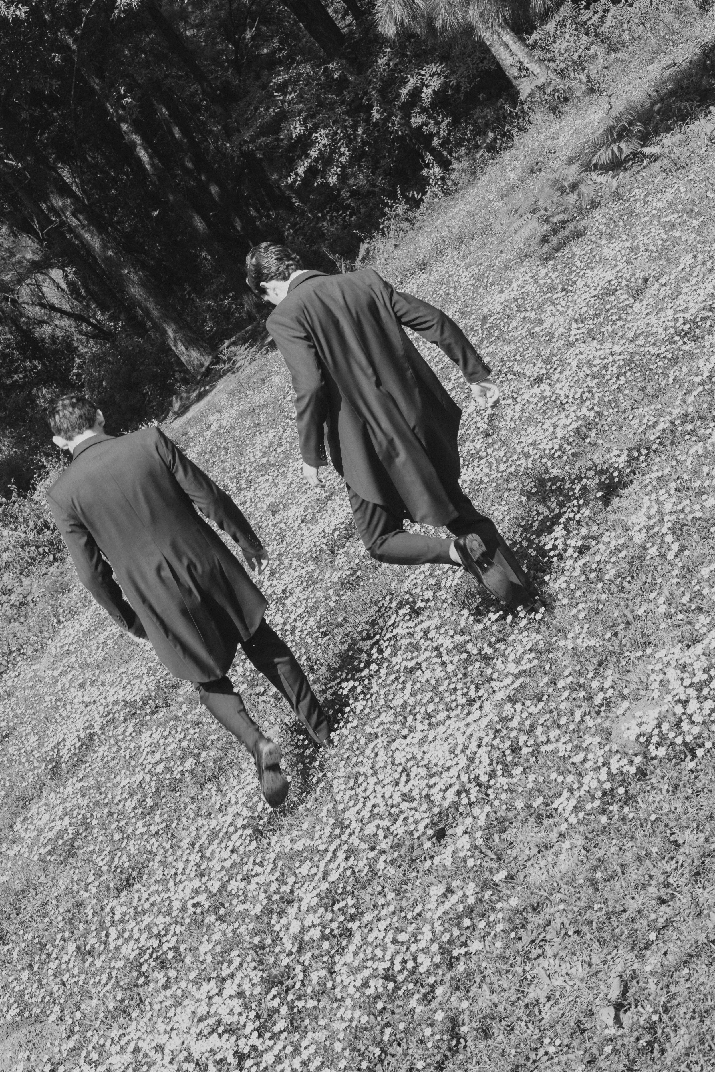 Dos hombres con trajes caminando por un sendero en un bosque en blanco y negro.