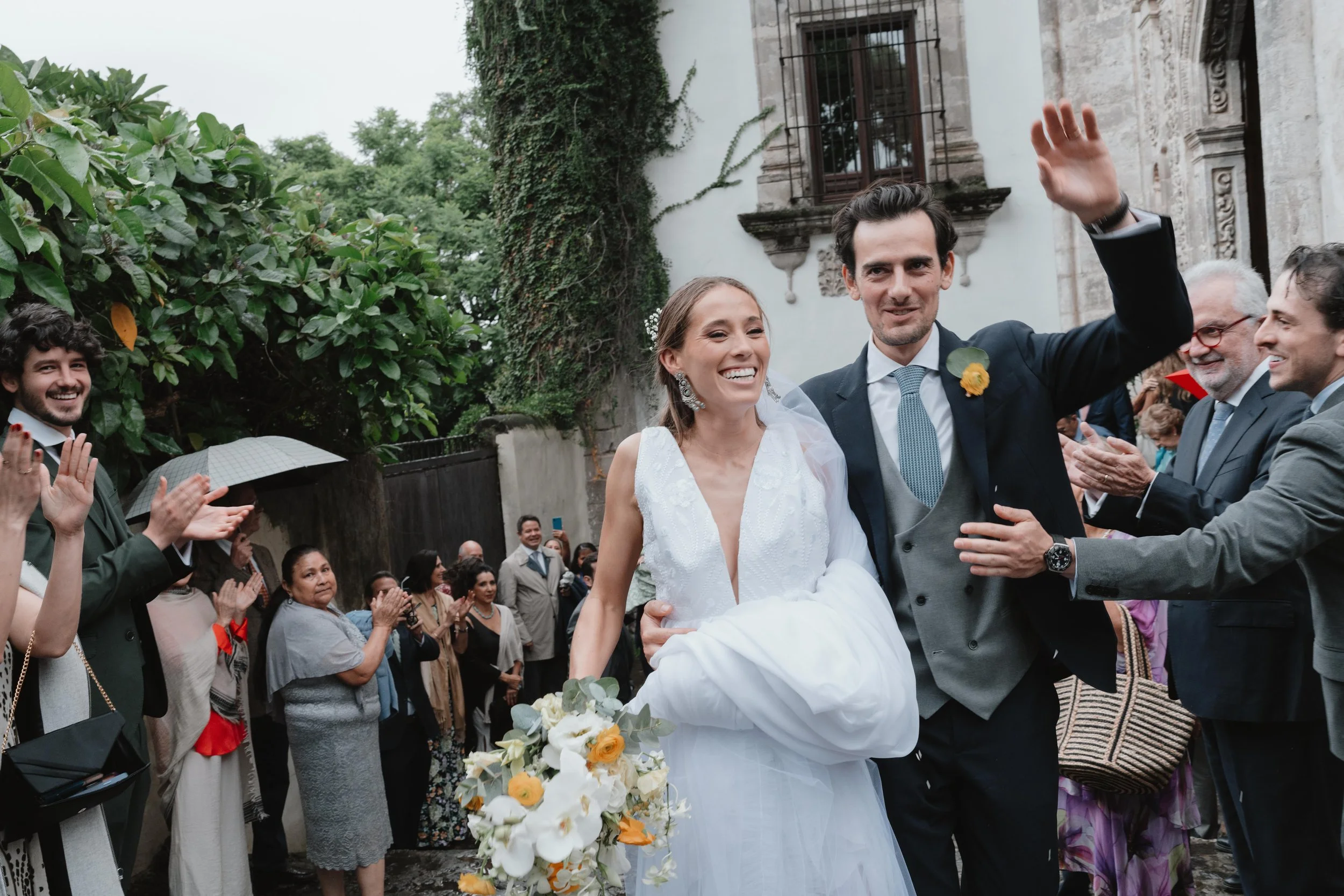 Pareja de recién casados en una ceremonia con invitados aplaudiendo y celebrando afuera de una iglesia antigua, rodeada de vegetación.