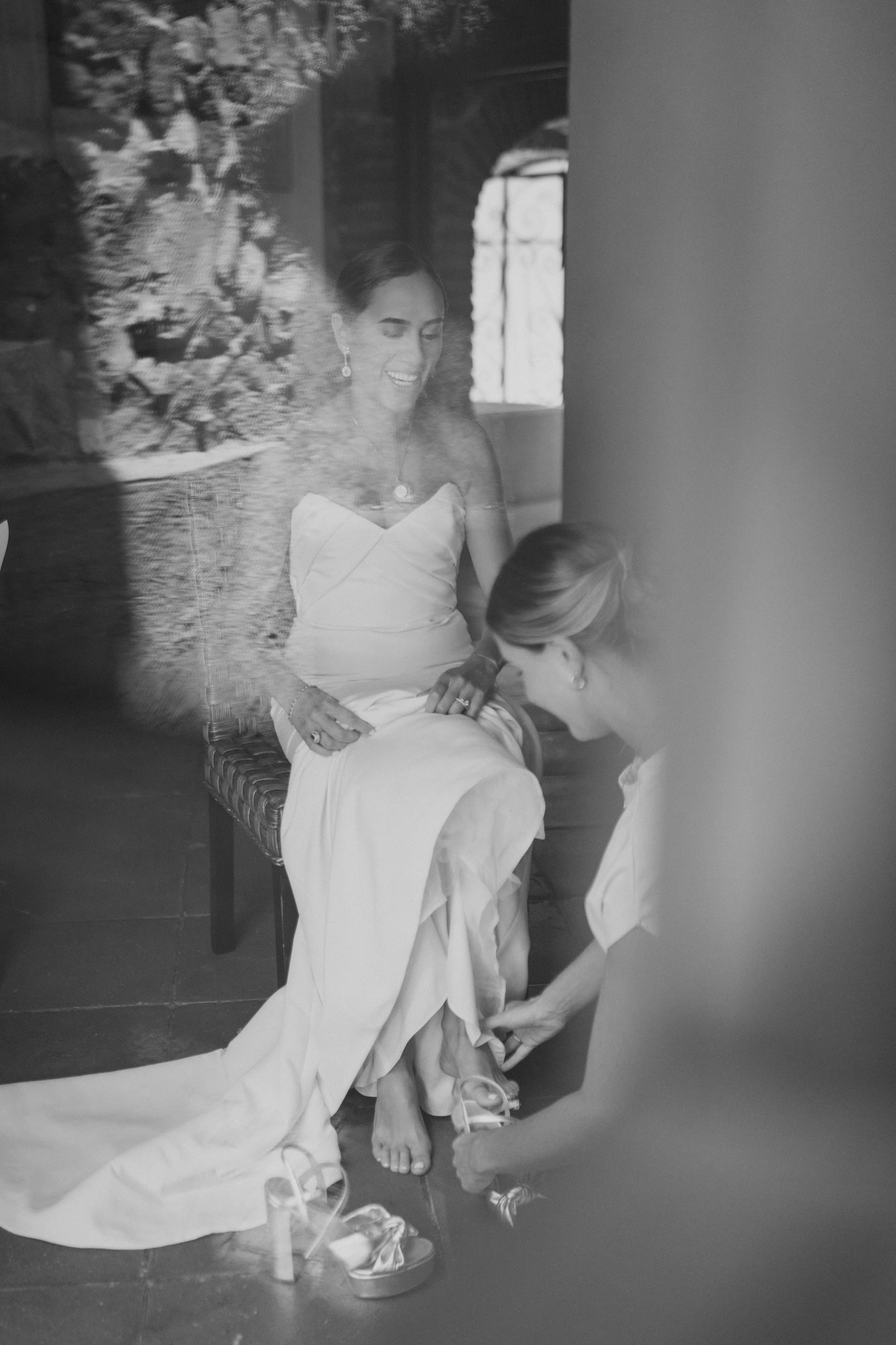 Mujer en vestido de novia siendo ayudada a ponerse los zapatos por otra mujer, vista a través de una ventana o puerta con reflejo.