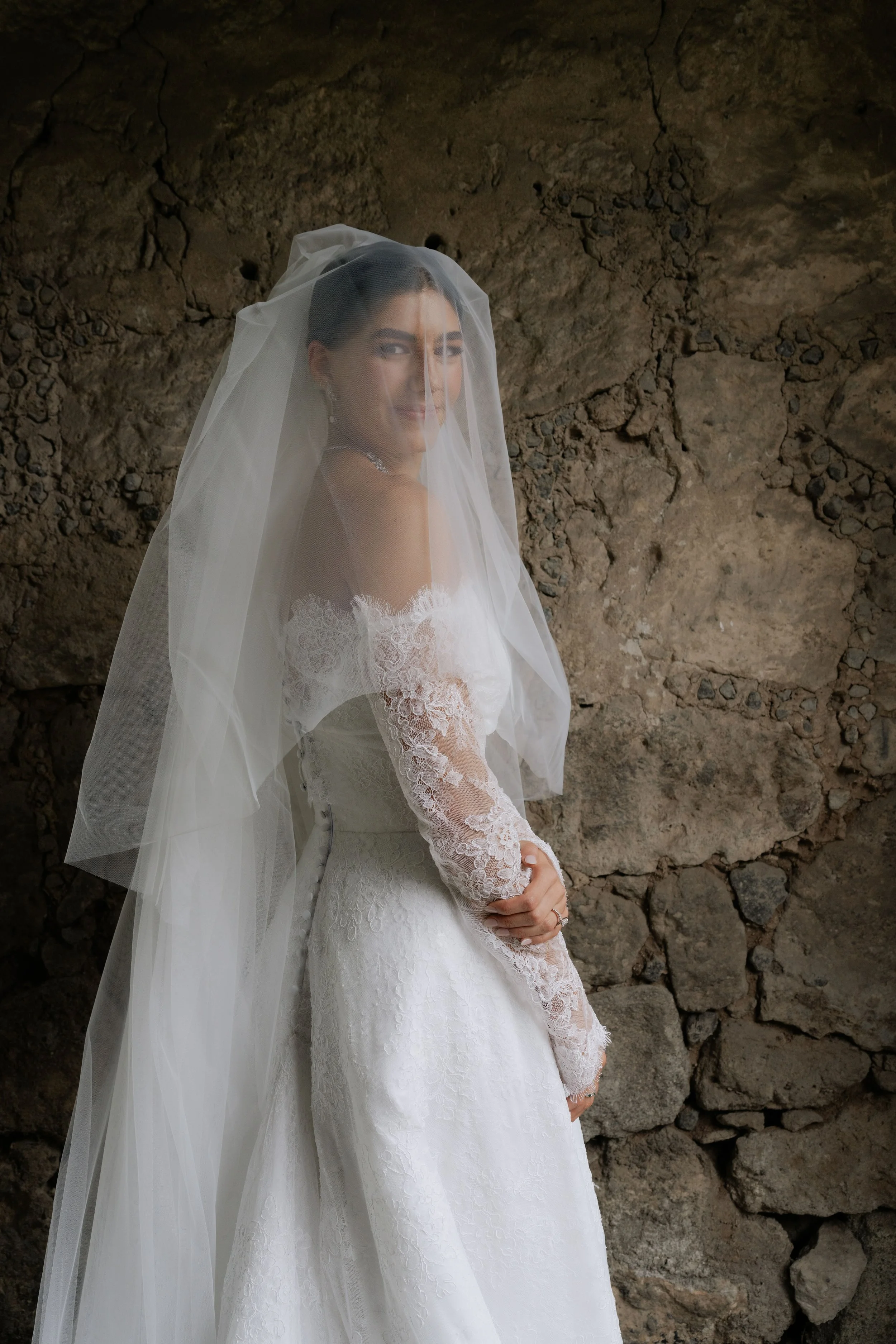 Mujer vestida con un vestido de novia blanco con encaje y velo, de pie junto a una pared de piedra, sonriendo.