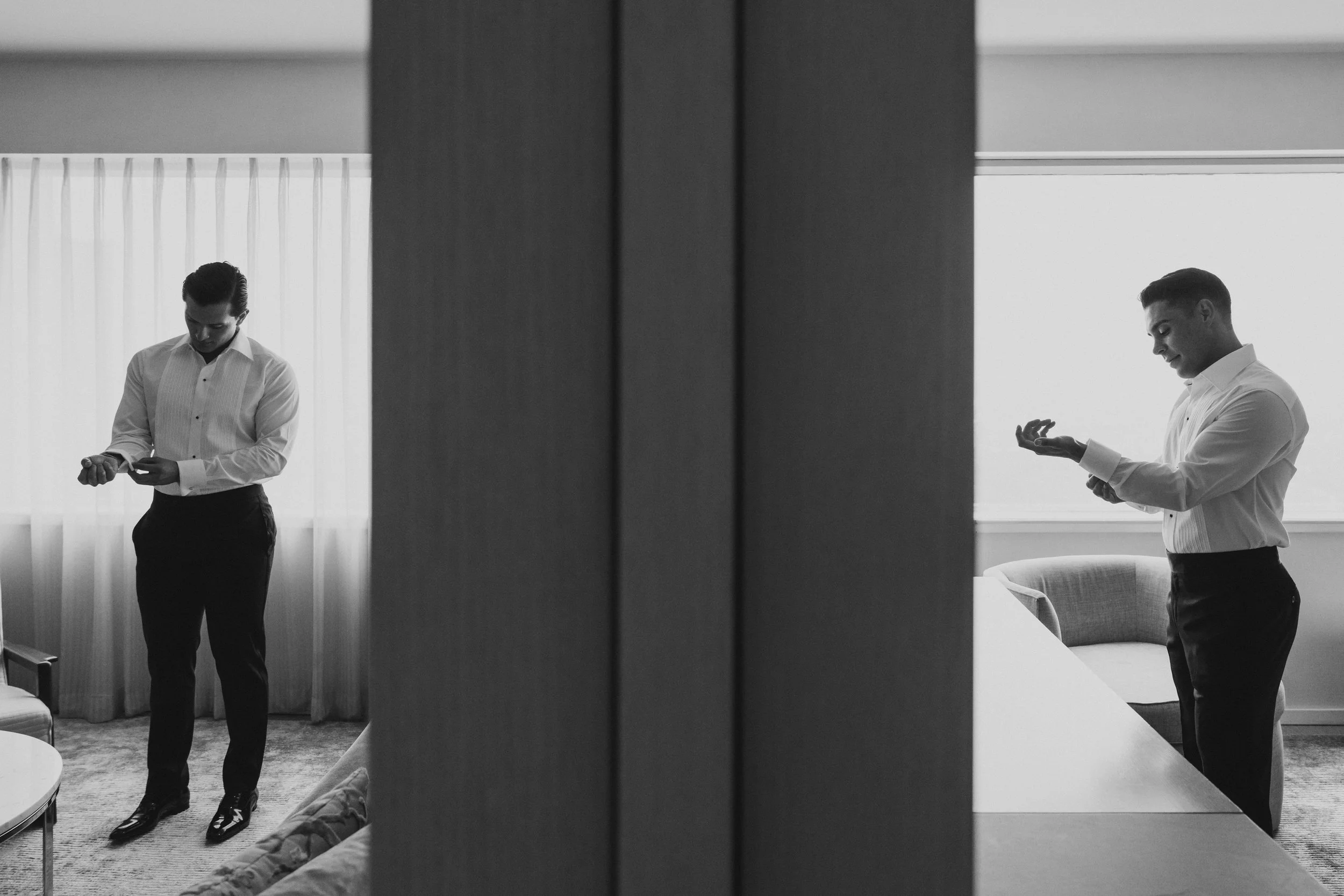 Imagen en blanco y negro de un hombre con camisa blanca y pantalones oscuros ajustándose las mangas en una habitación con cortinas blancas y una ventana que deja pasar luz natural.