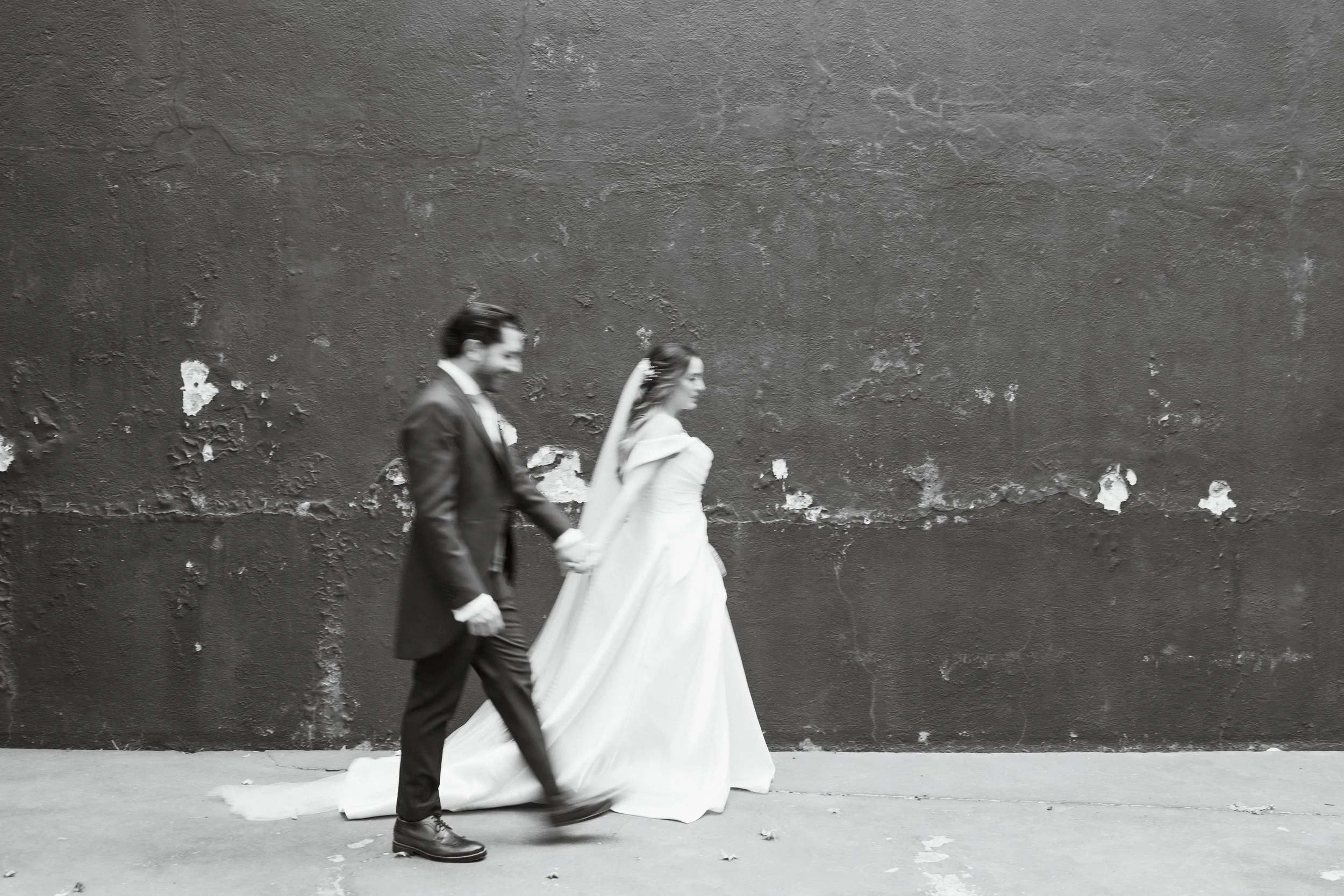 Pareja casándose, la mujer con vestido de novia y el hombre con traje, caminando juntos de la mano frente a una pared de textura áspera.