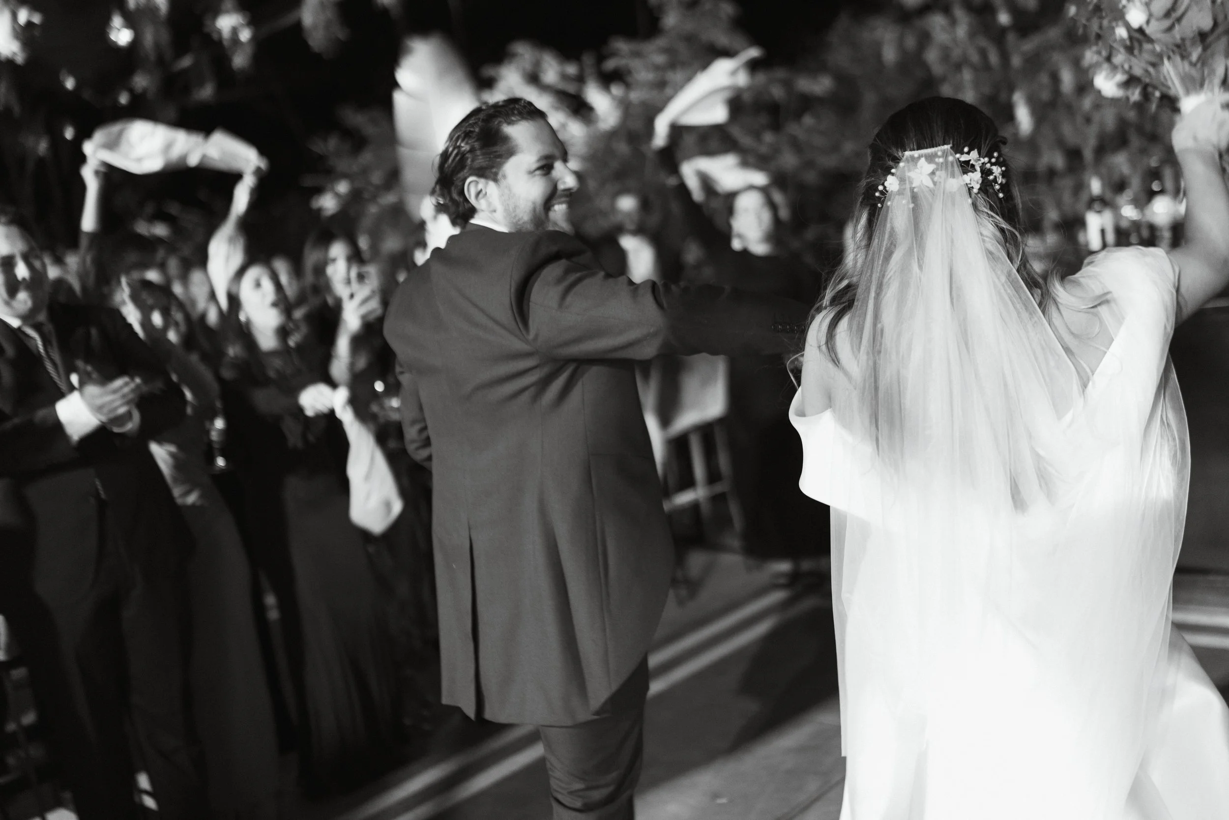Pareja bailando en una boda, con invitados observando y tomando fotos en el fondo, en un ambiente festivo.