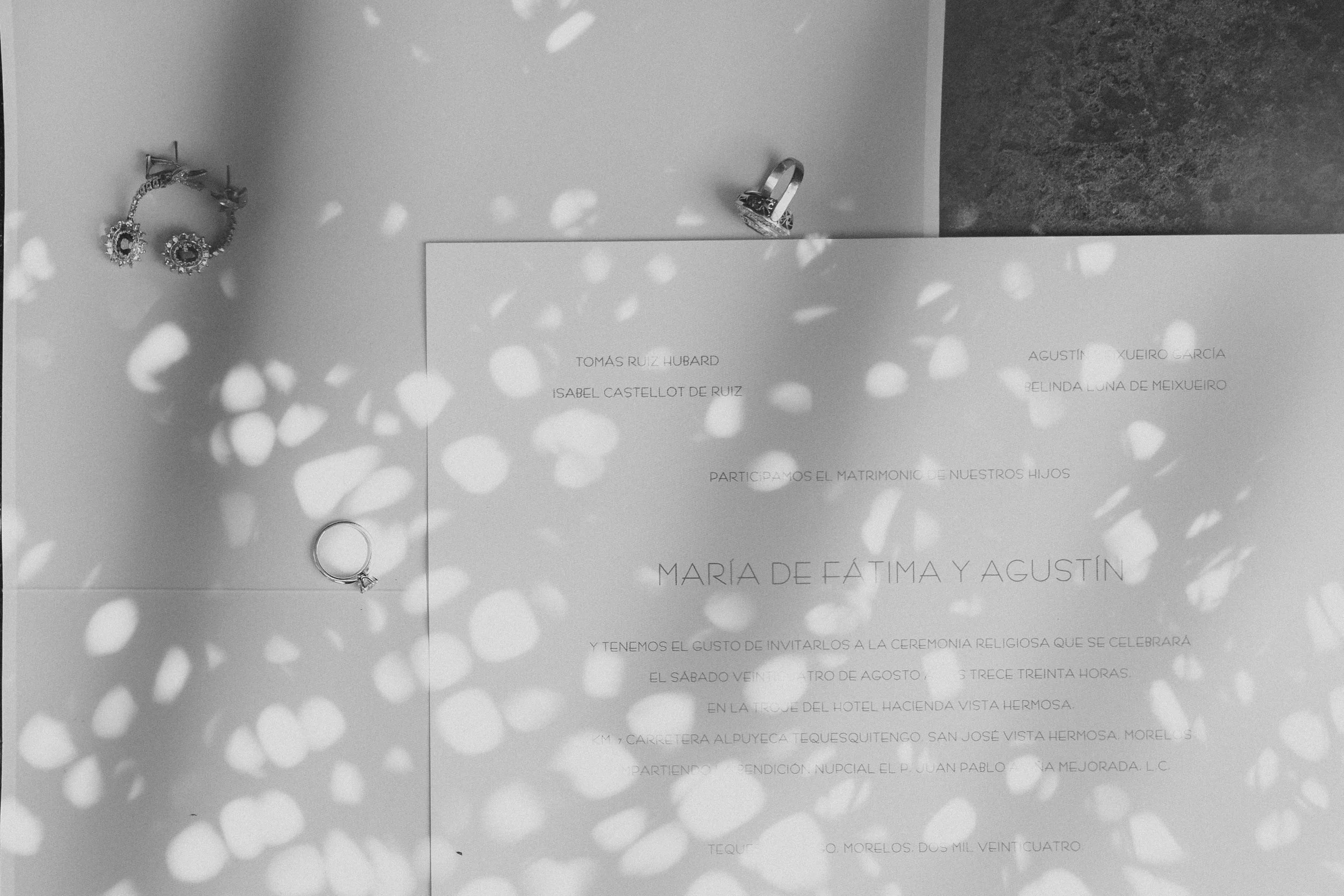 Invitación de boda con texto en español, junto a joyería y anillo. La invitación tiene sombras de pétalos o luces en el fondo.