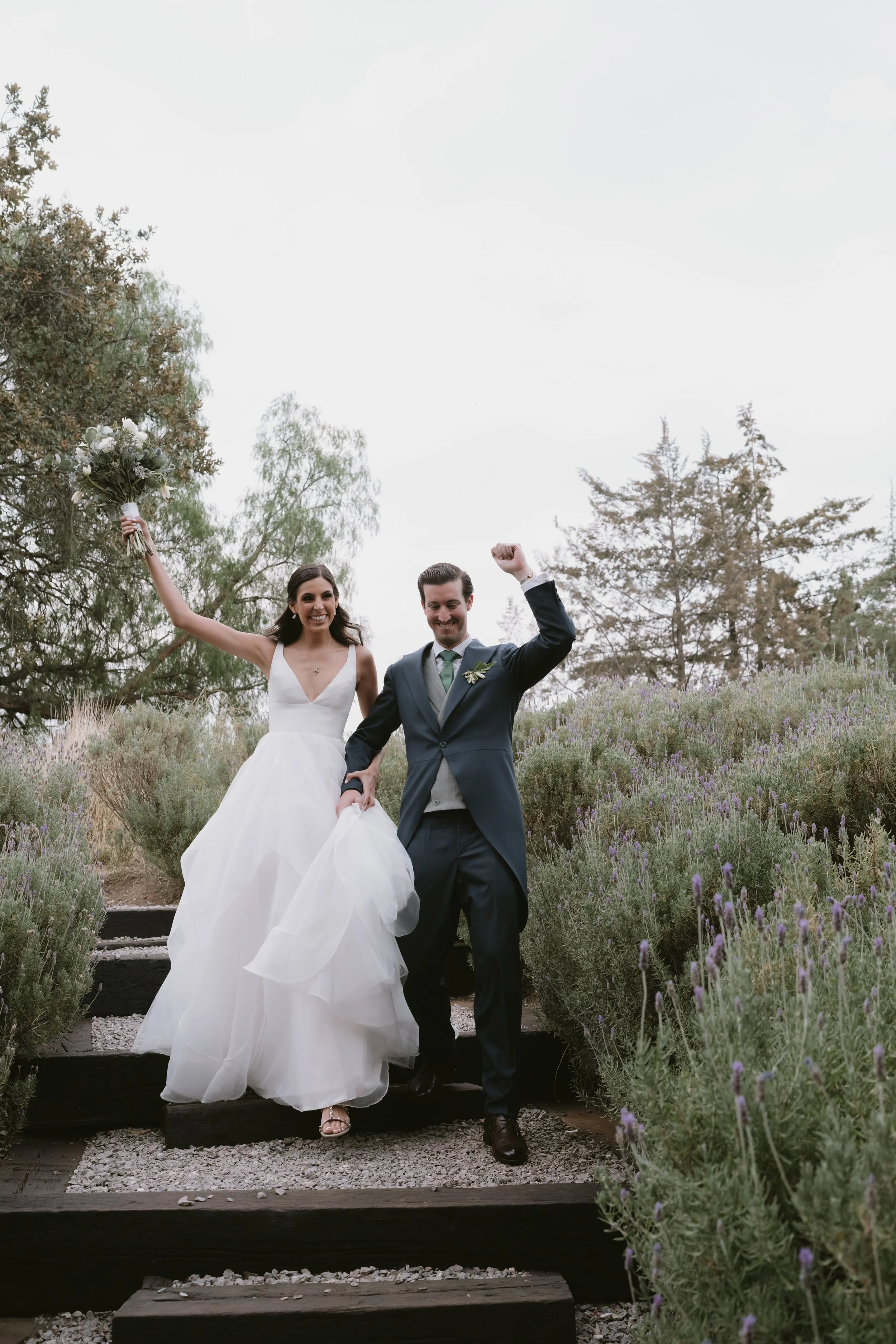 Pareja de recién casados celebrando, ella con vestido blanco y bolso de flores, él con traje oscuro y corbata verde, en un jardín con lavandas y árboles.
