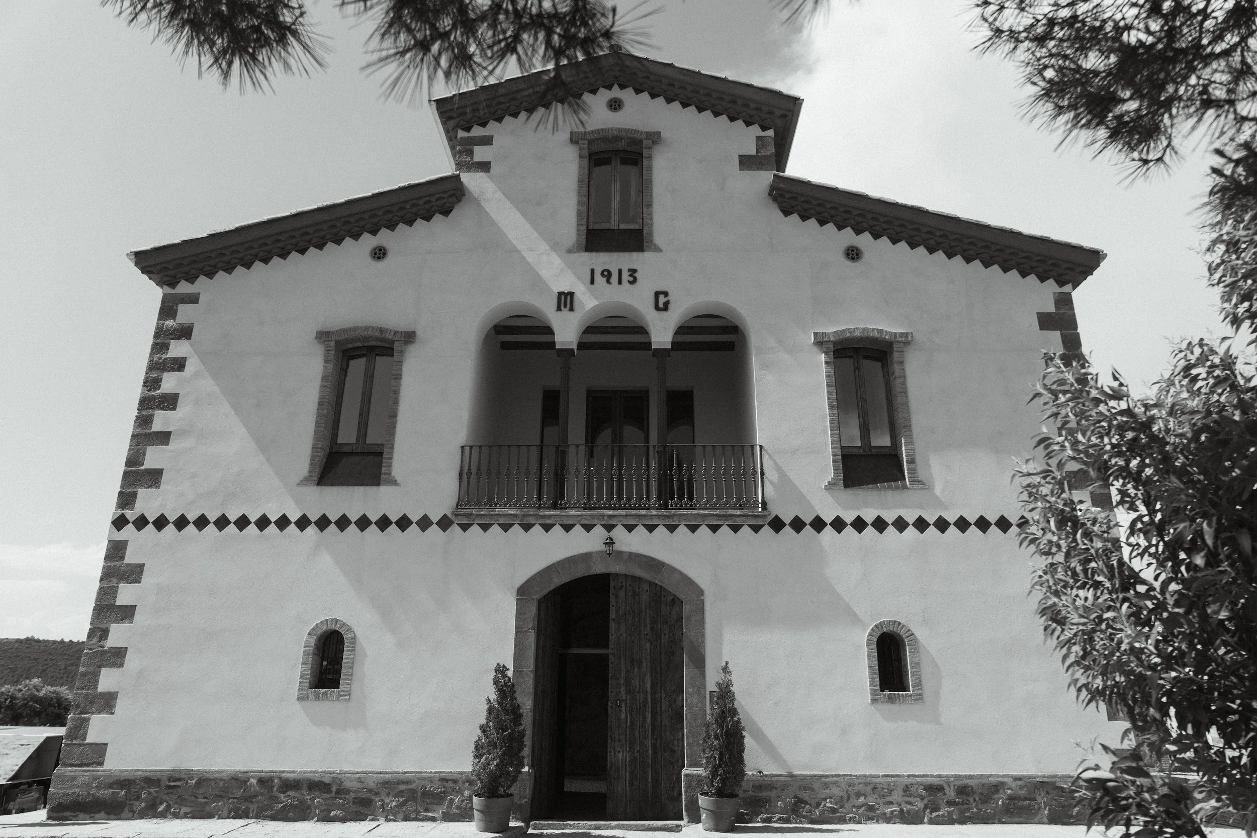 Casa antigua de estilo europeo con estructura de piedra y paredes blancas, ventanas de madera y balcón con reja, rodeada de árboles y plantas.