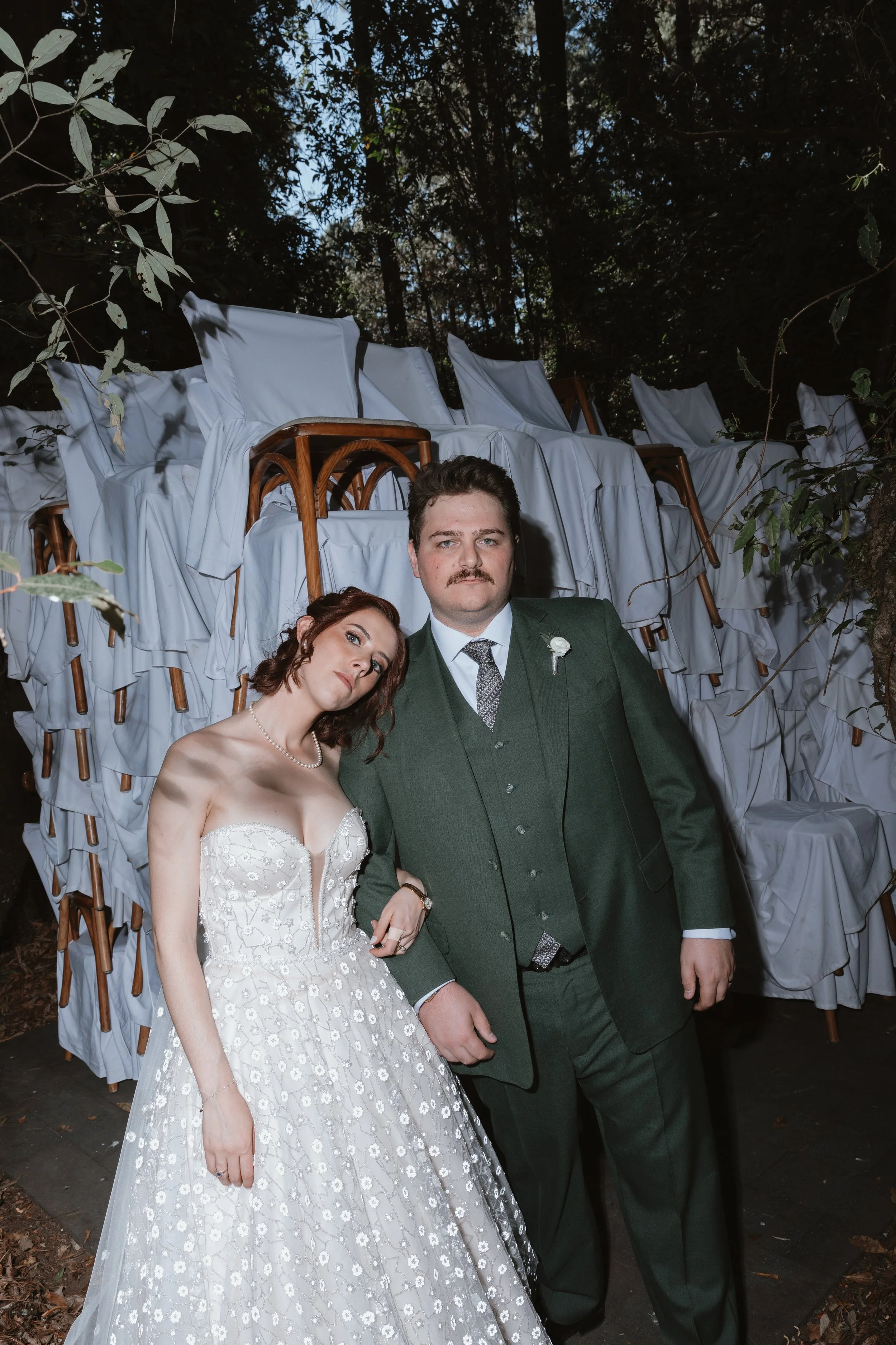 Una pareja en vestido y traje formal de boda, posando en un bosque con sillas apiladas y cubiertas con sábanas blancas en el fondo.