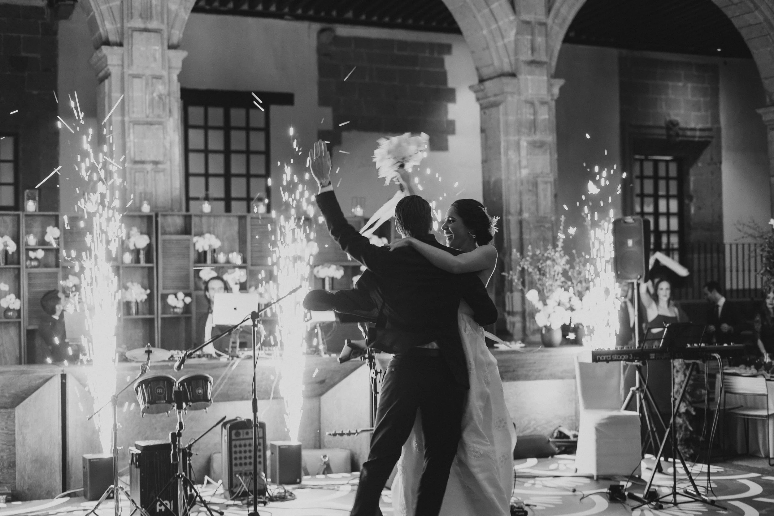 Pareja de recién casados bailando en su boda con fuegos artificiales y música en vivo en segundo plano, en un lugar elegante y decorado con flores.