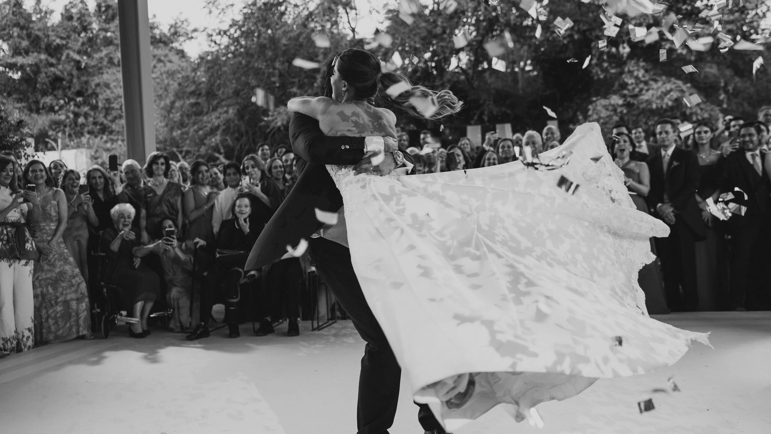 Pareja bailando en una boda, rodeada de invitados, en un ambiente al aire libre, mientras confeti cae del cielo.