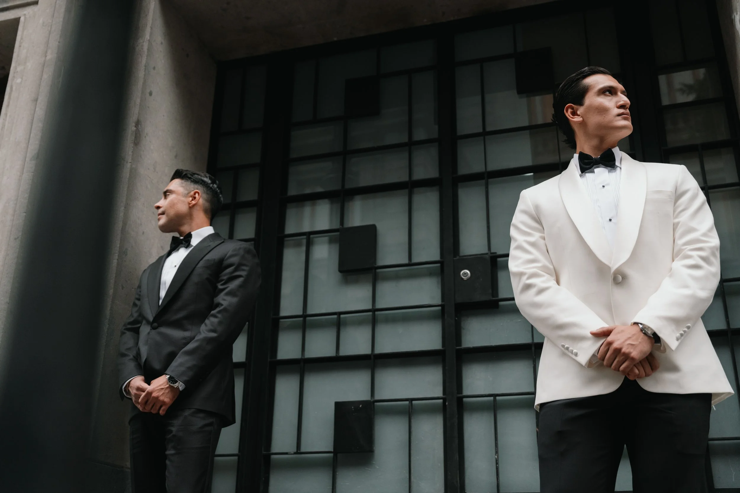 Dos hombres en trajes formales de esmoquin, uno en negro y otro en blanco, de pie frente a una estructura de ventanas modernas en una construcción de ladrillo y cemento, en actitud seria y formal.