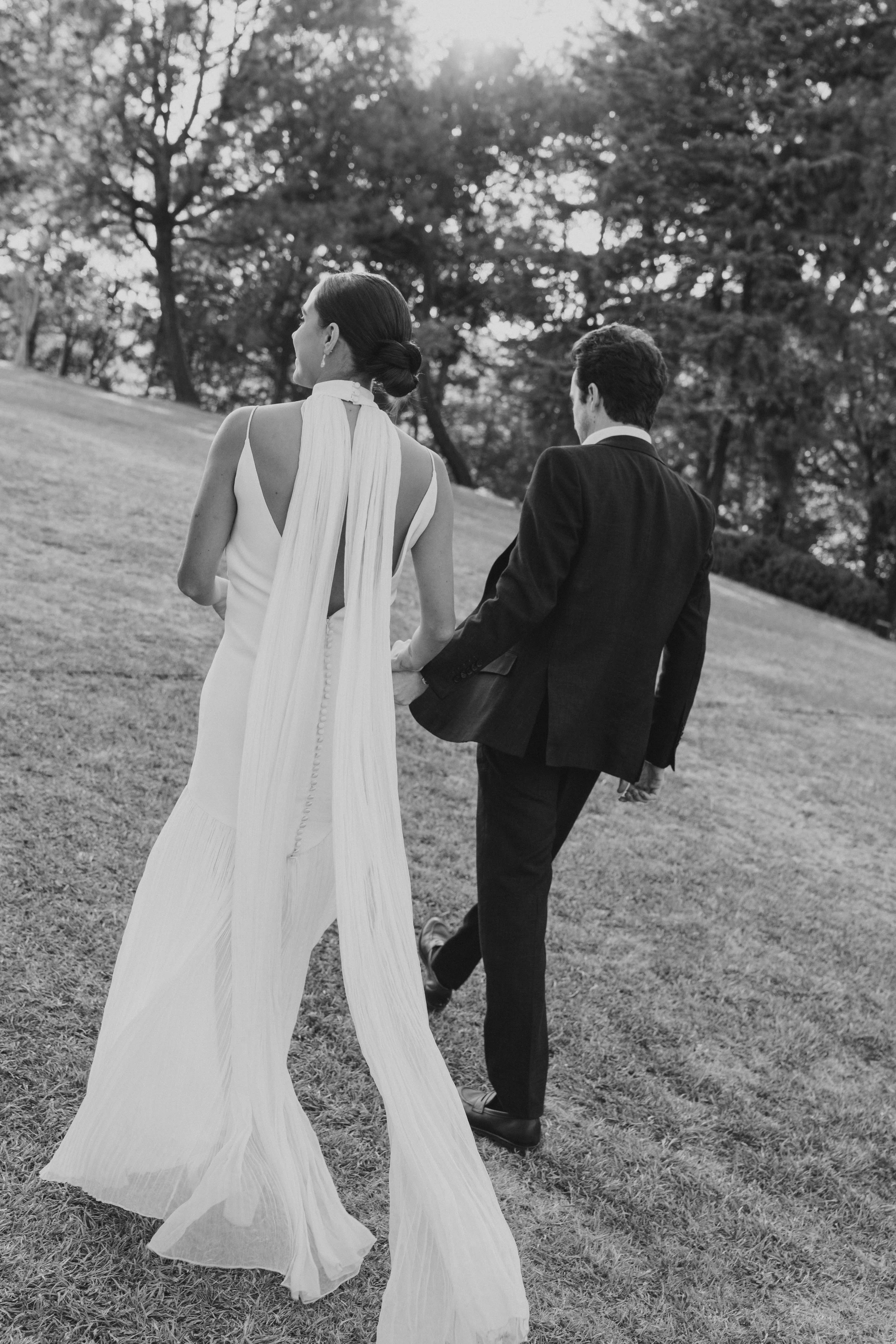 Pareja vestida formalmente, con la mujer en un vestido largo y el hombre en un saco, caminando por un parque con árboles y césped.