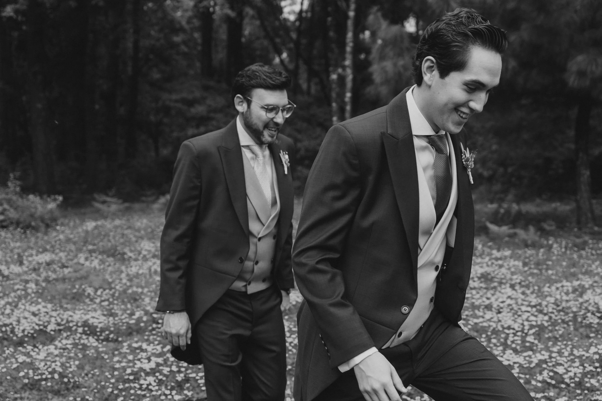 Dos hombres vestindo trajes formales caminando por un Bosque, uno sonriente en primer plano y el otro con barba y gafas en segundo plano, en un entorno natural