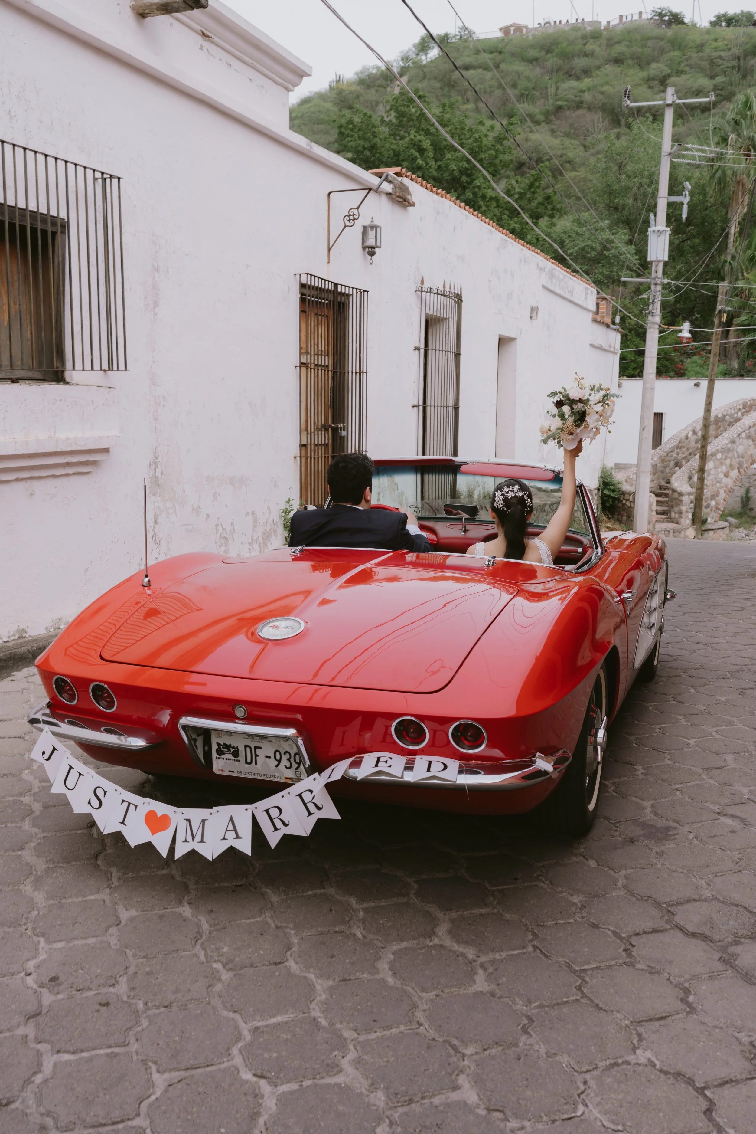 Pareja de novios en un coche clásico rojo, decorado con una pancarta que dice "JUST MARRIED", en un camino de piedra, con paredes blancas y colinas verdes de fondo.