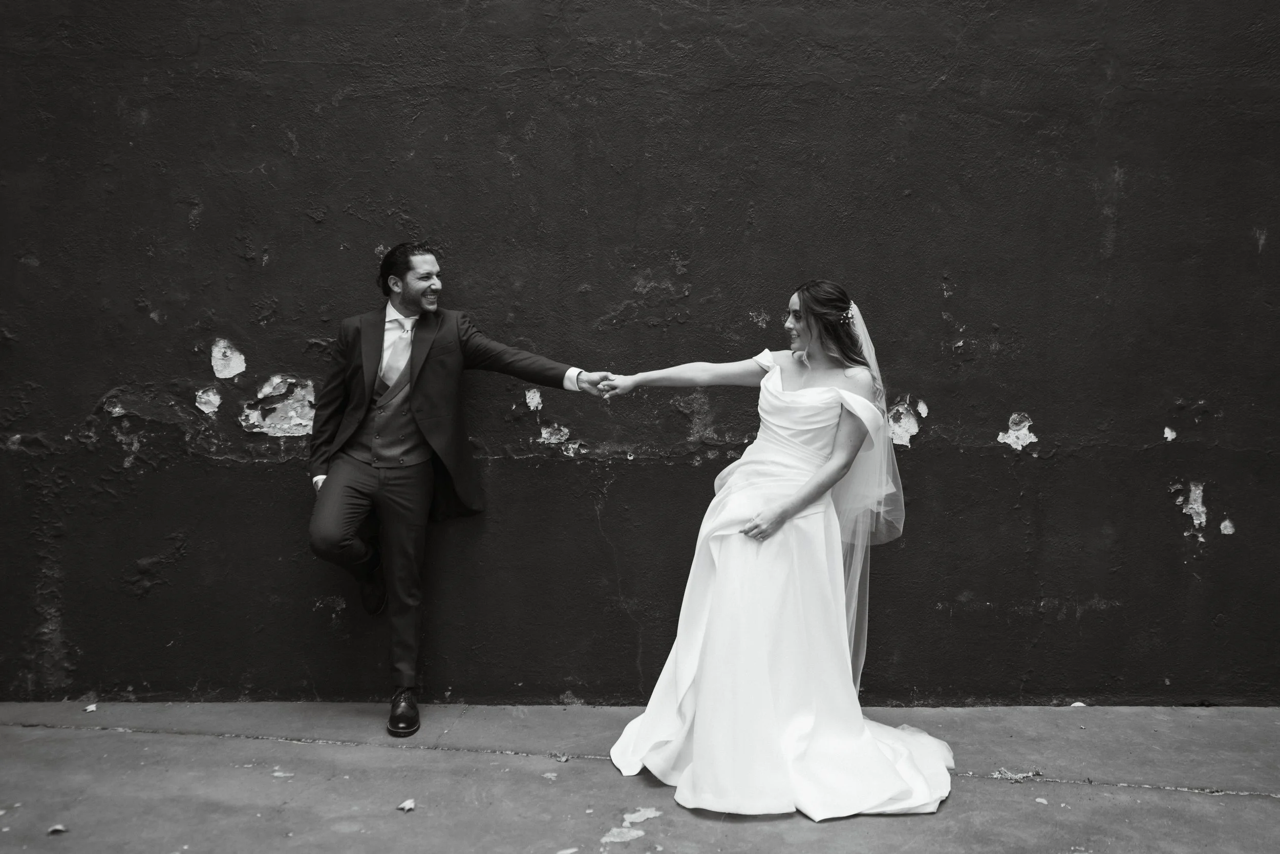 Una pareja de recién casados, vestido de novio y novia, se toman de las manos frente a una pared oscura, sonriendo y disfrutando su momento.