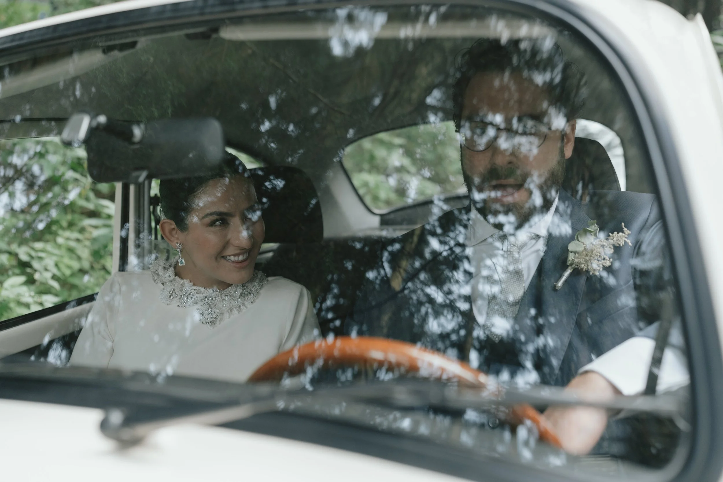Una pareja vestida de boda dentro de un automóvil, la mujer sonríe y lleva un vestido blanco con joyería, el hombre viste un traje con boutonnière, ambos parecen ser novios.