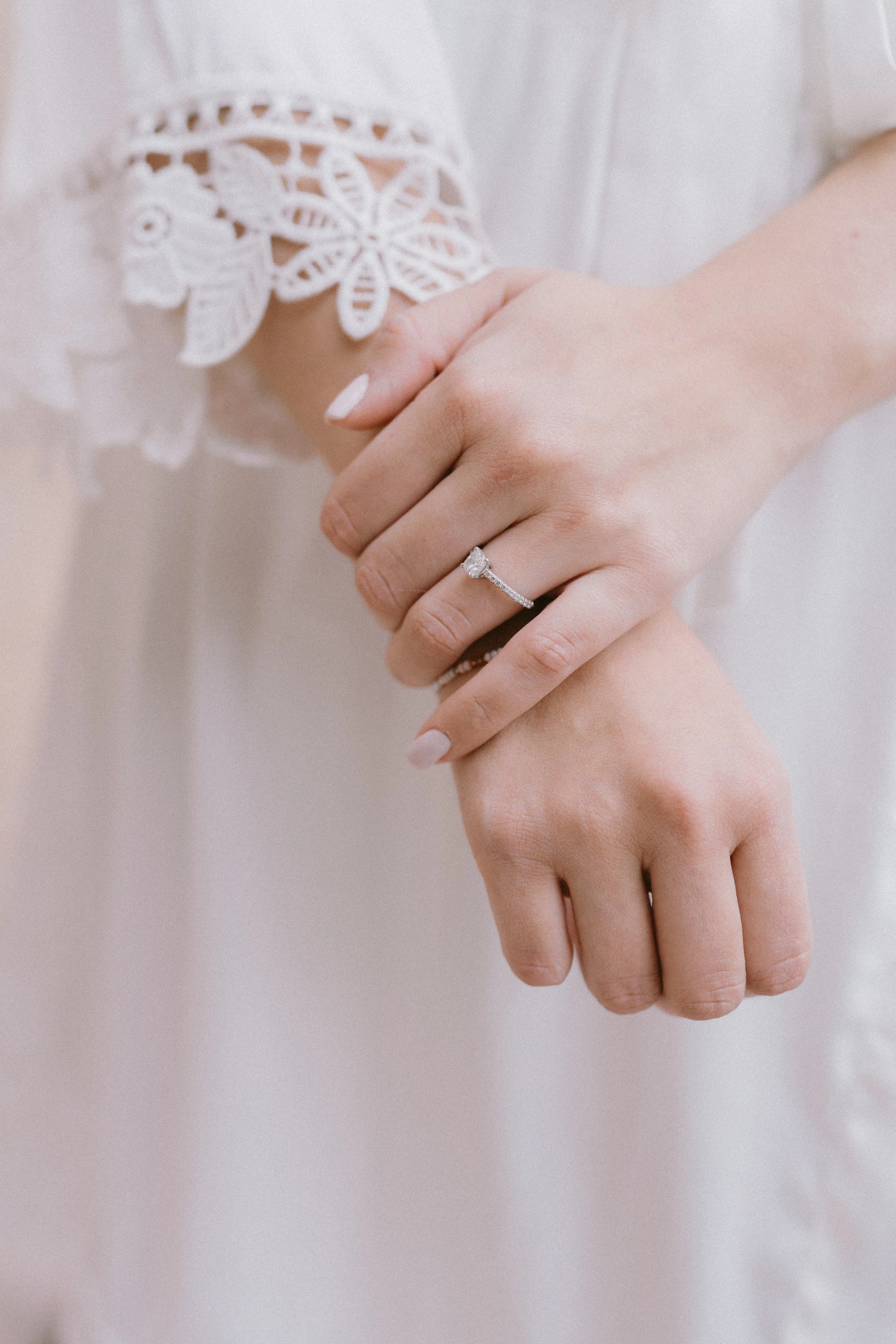 Manos entrelazadas mostrando un anillo de compromiso con diamante en un dedo y una blusa blanca con detalles de encaje.