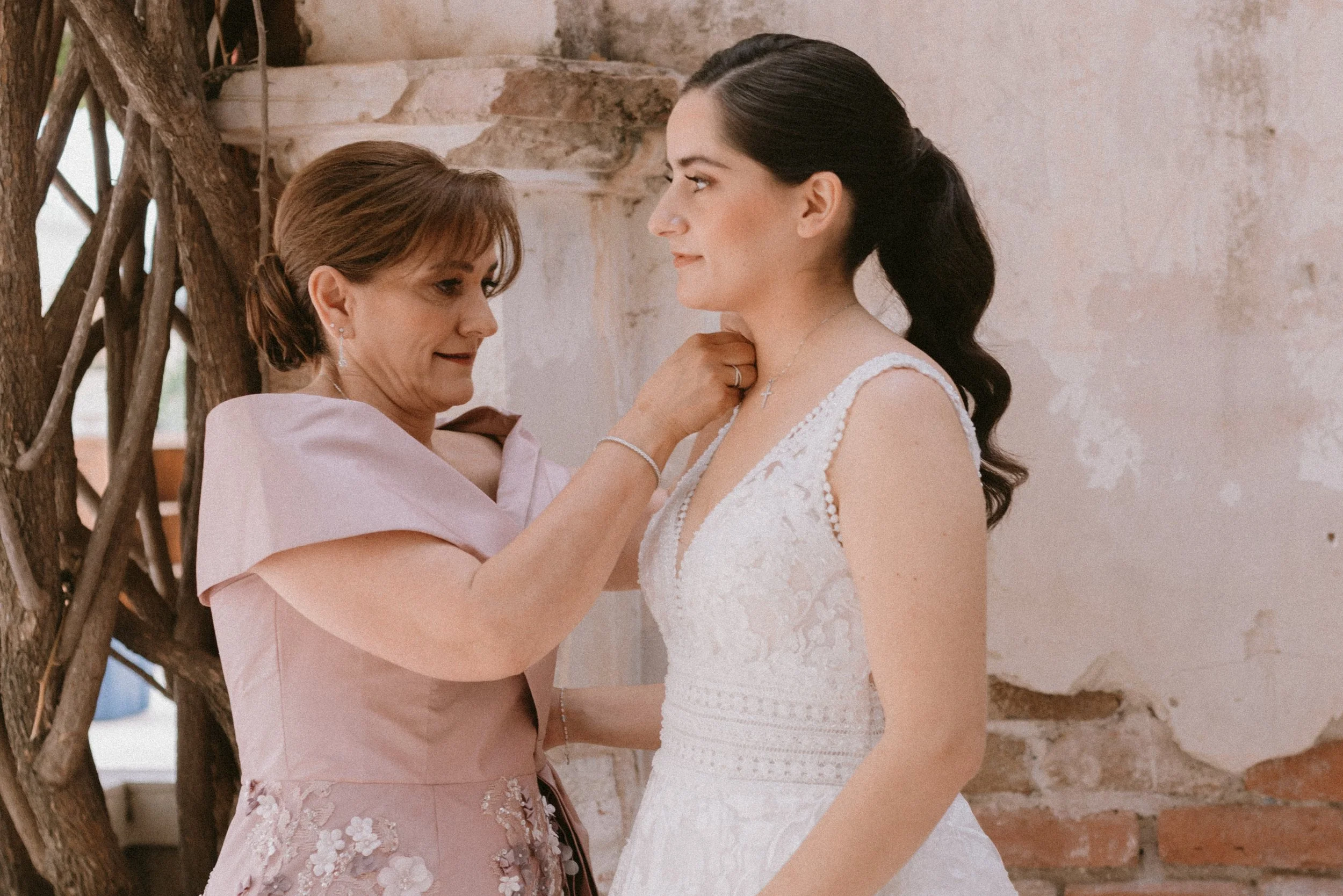 Una mujer mayor ayuda a una novia a colocarse un collar antes de la ceremonia de boda en un lugar con paredes de ladrillo y decoración natural.