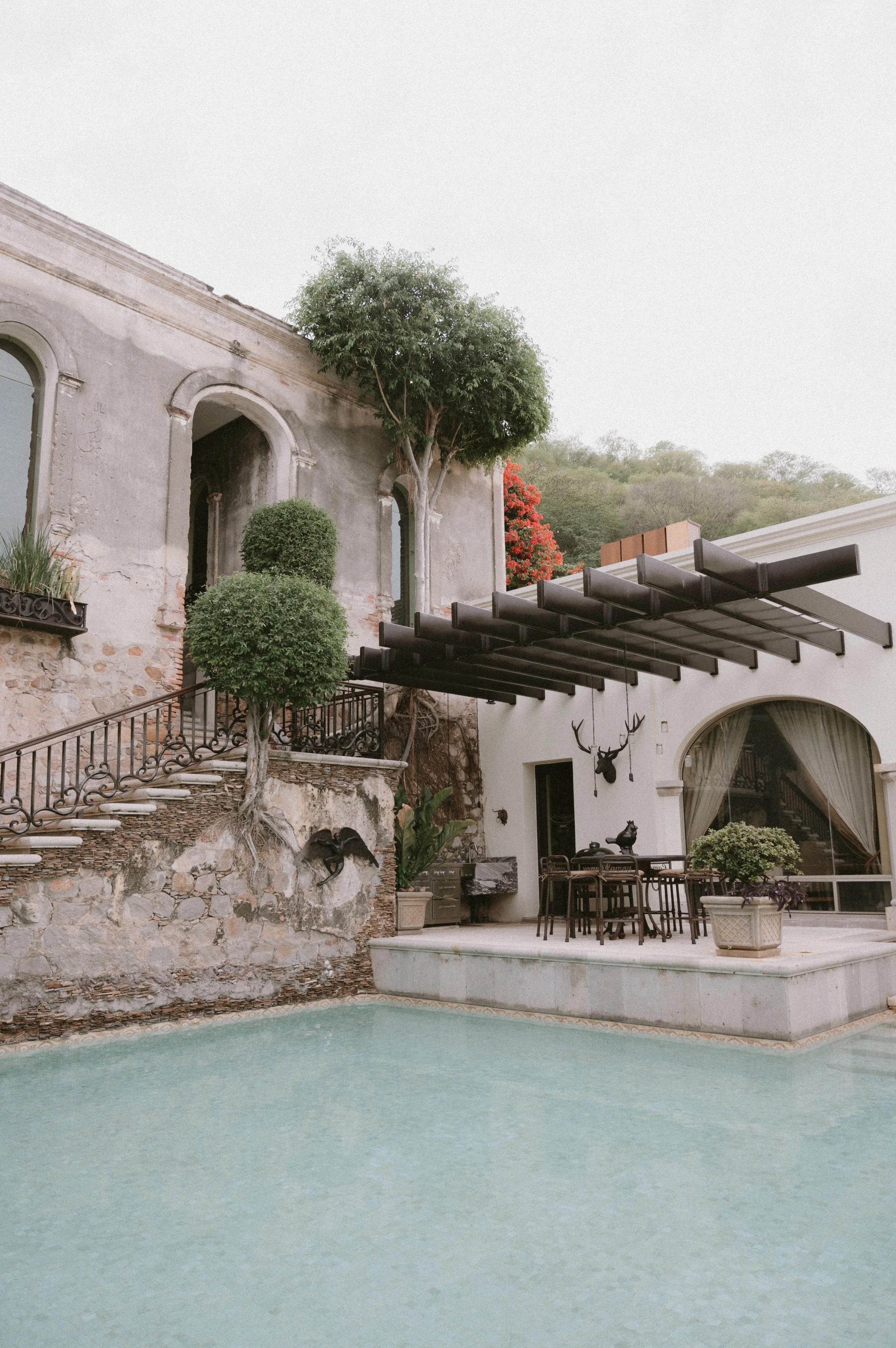 Casa con muro de piedra y ventanas arqueadas, área de patio con piscina, árboles podados en forma de bola, mesa con sillas, decoración con cabeza de ciervo y animales decorativos, en un ambiente tranquilo y natural.