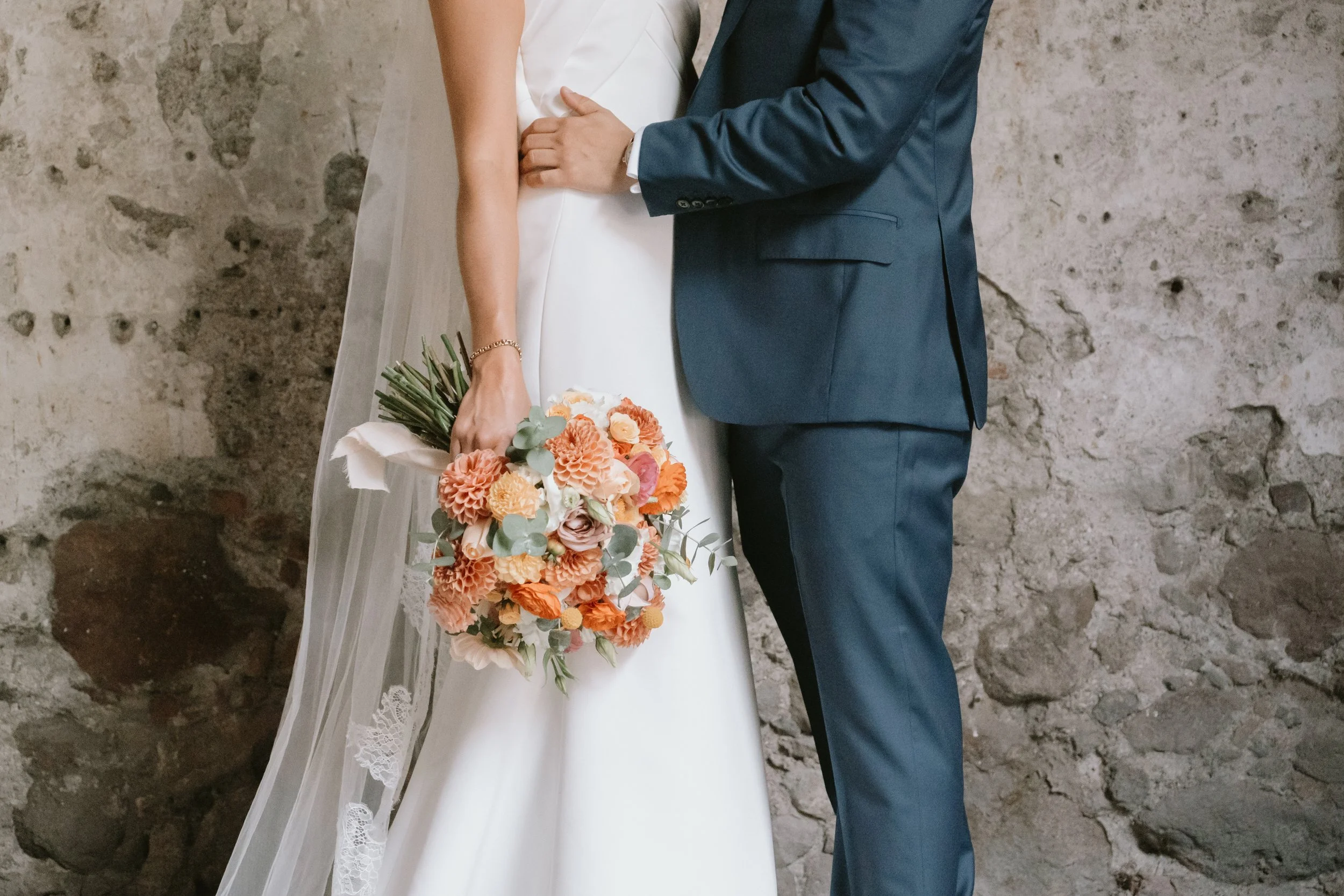 Pareja de novios en una boda, con una mujer sosteniendo un ramo de flores con tonos anaranjados y verdes, y un hombre vestido con saco azul. Fondo de pared de piedra.