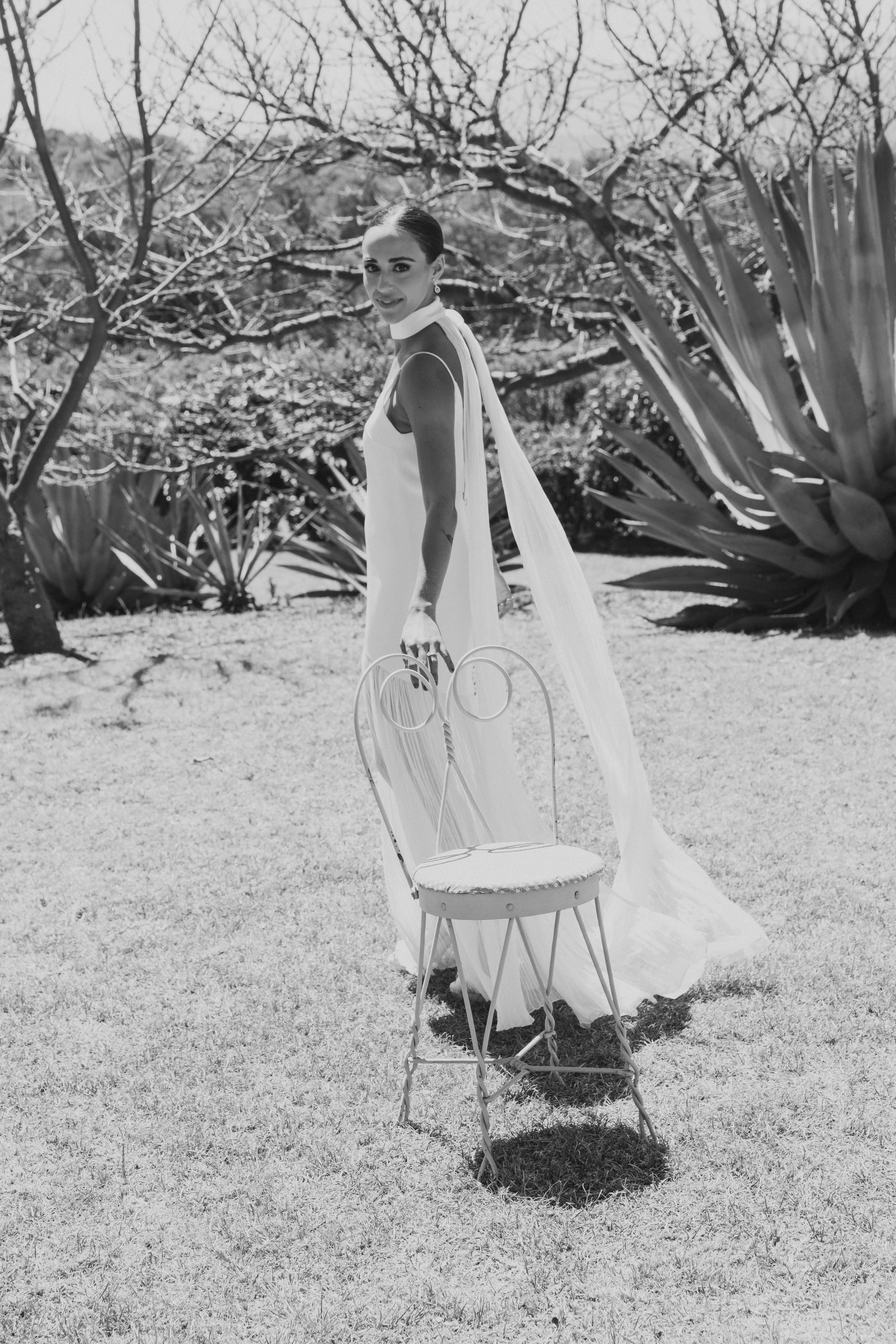 Mujer vestida de blanco en un jardín con plantas grandes y árboles sin hojas, en un día soleado.